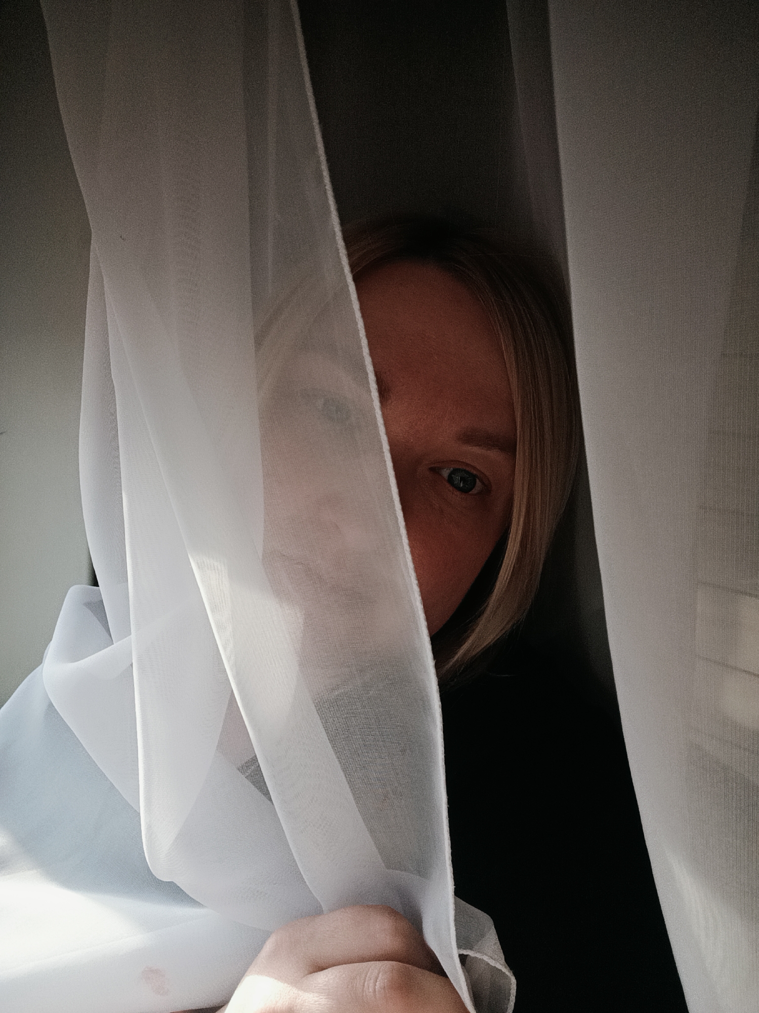 A woman is partially visible, peeking from behind sheer white fabric, likely a curtain, in an indoor setting in Chaikovsky, Russia. Her right eye, part of her nose and forehead, and strands of her blonde or light brown hair are discernible. Her hand holds the sheer material at the bottom of the frame. The fabric diffuses the light, suggesting daylight, possibly creating a soft, ethereal glow. The area behind the woman appears dark, providing a stark contrast to the illuminated fabric and her skin tone. Her visible eye is open and appears to be looking directly forward. The scene evokes a sense of intimacy, mystery, or quiet observation. No text is visible in the image.