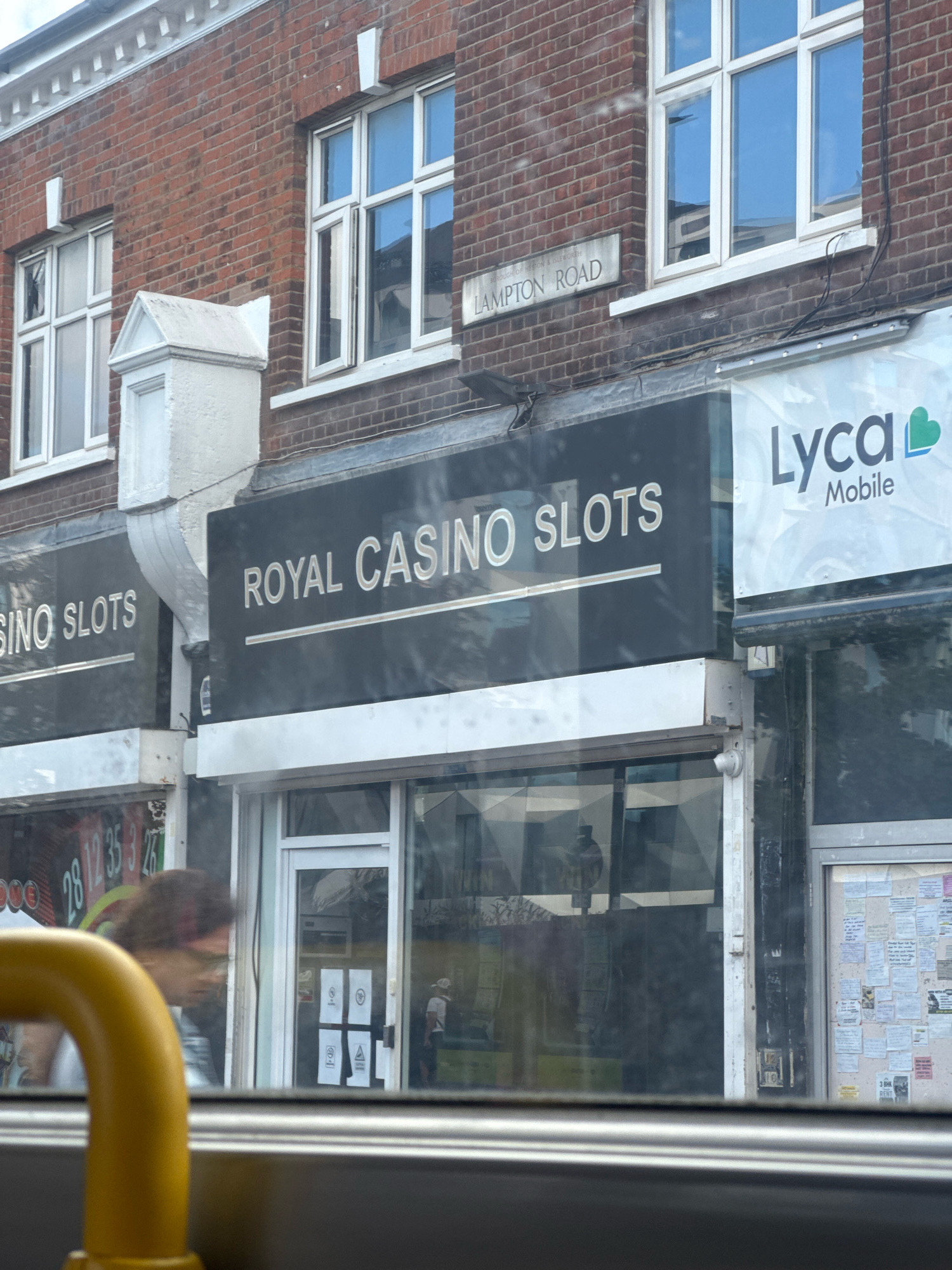 The image shows a street scene in Hounslow, London, United Kingdom. A red brick building with white-trimmed windows is visible. A street sign reads "LAMPTON ROAD". Below the street sign, a dark-colored awning displays the text "ROYAL CASINO SLOTS" in white lettering. To the right of the casino awning, another awning displays the logo and text "Lyca Mobile". The ground floor of the building features large glass windows, through which various advertisements and signs are visible. A person with dark, curly hair is seen through the window, appearing to be walking. A yellow handrail is in the foreground, obstructing a portion of the view of the street level. The overall scene depicts a commercial area with multiple businesses.
