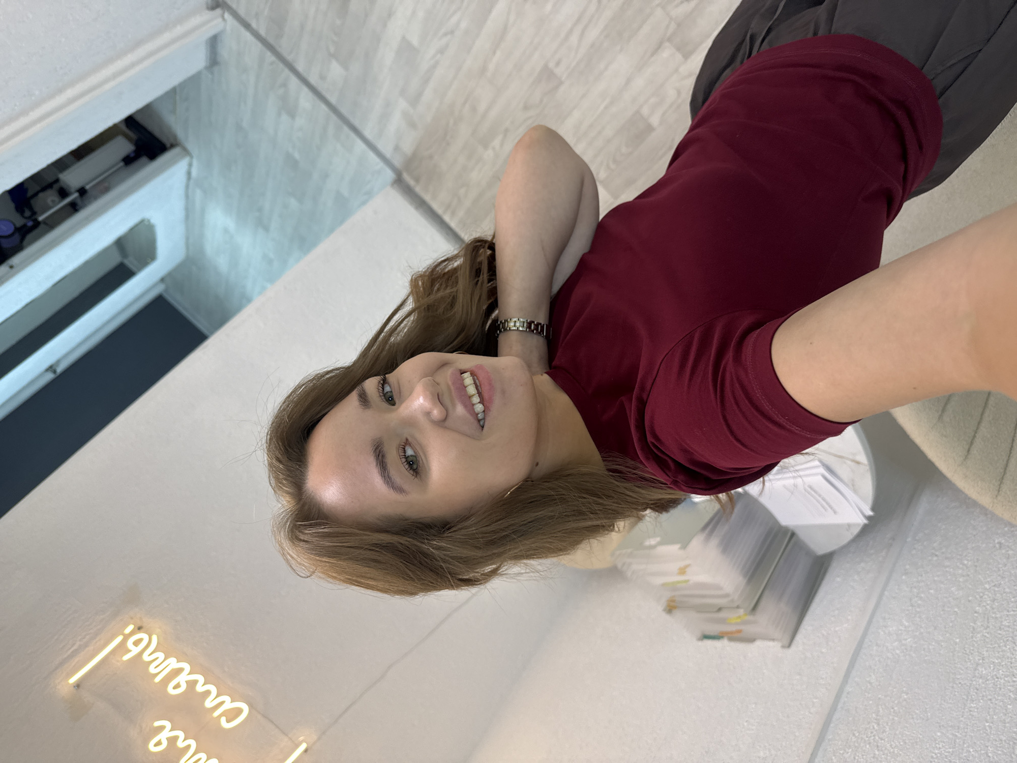 In a dimly lit room, a young woman with shoulder-length brown hair, styled casually, takes a selfie. She is wearing a maroon t-shirt and dark pants. Her right arm is extended towards the camera, and her left hand is touching her neck. She has a bright smile, with her teeth visible. Behind her, a stack of papers is on a surface, and to her left, a neon sign with Cyrillic lettering glows in yellow. The room's flooring appears to be light-colored wood, and a doorway is visible in the background, revealing a vacuum cleaner and a white wall. The overall impression is of an informal setting, possibly a home or a personal workspace, located in the city of Chaikovsky, Russia, given the specific neon sign which translates to "Come shine!" or "Be bright!".