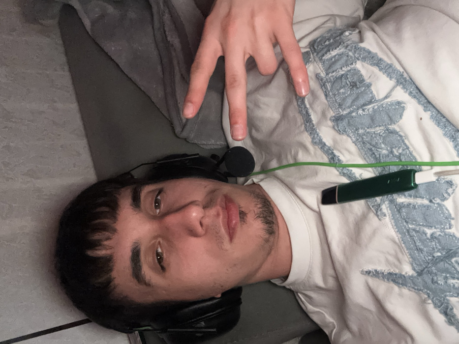 The scene is set indoors, within the city of Vetrino, Bulgaria. A single male individual is depicted in a medium close-up, facing the camera directly. He possesses dark, short hair styled with a straight fringe across the forehead and light stubble on his chin and upper lip forming a goatee. He wears black over-ear headphones with an attached microphone boom, which is positioned to his left. A thin, bright green cable extends downwards from the headphones across his chest. He is dressed in a light-colored (possibly off-white or cream) long-sleeved shirt featuring a distressed light blue fabric design on the front. His left hand is raised to chest level, forming a gesture with the index and pinky fingers extended and the thumb, middle, and ring fingers curled. Resting on his shirt is a dark green, pen-shaped device, likely an e-cigarette or vape pen, which is connected to a short white charging cable. This white cable is draped over the green headphone cable. The individual appears to be reclining against a grey upholstered surface, possibly a sofa or bed, with a grey textured wall visible in the upper background.
