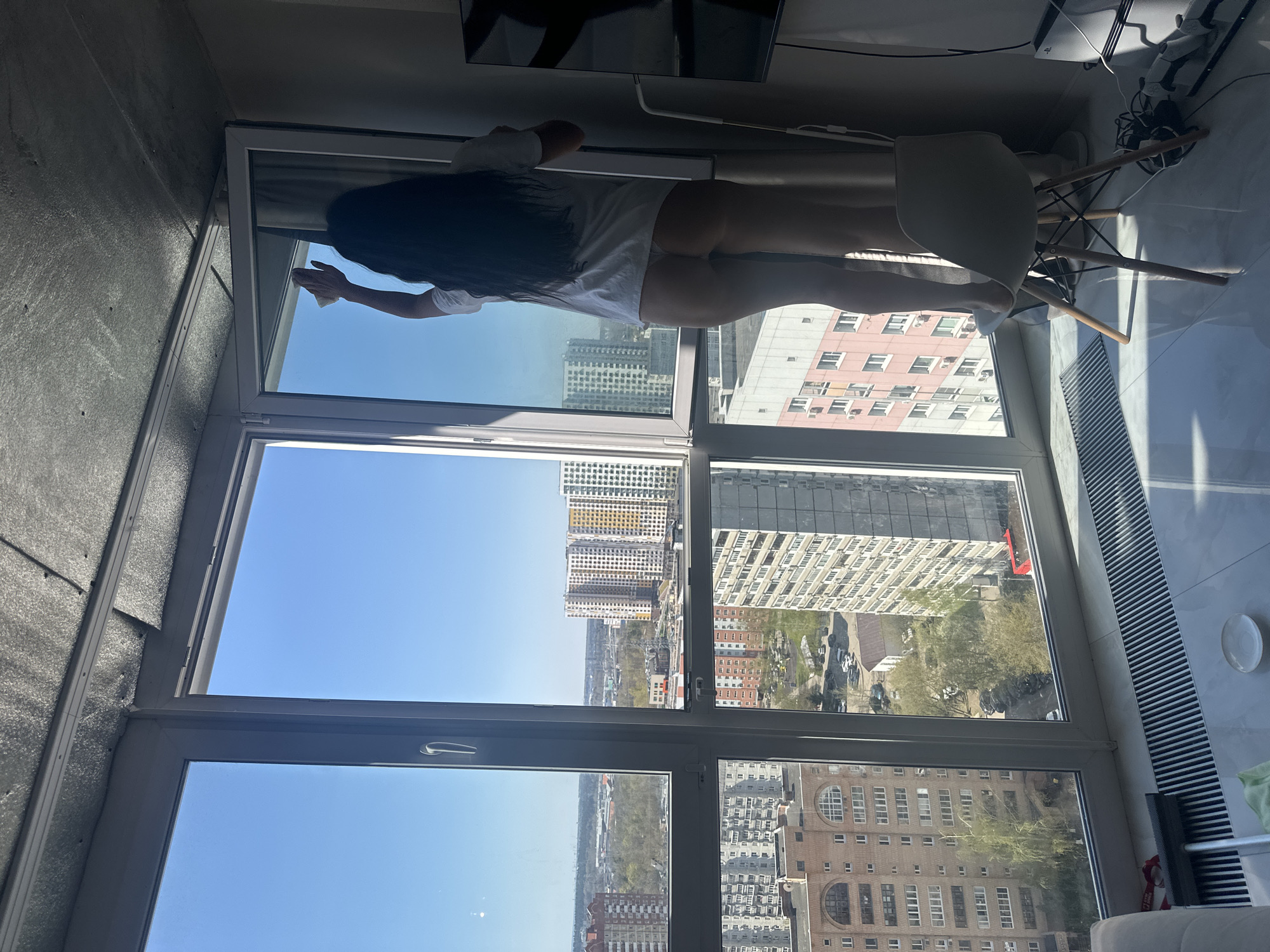 A photograph captured from inside an apartment in what is presumed to be Gavrilkovo, Russia, showcases an individual standing on a chair and cleaning a window, through which a cityscape unfolds under a clear blue sky. The individual, viewed from the back, has long, dark hair, is wearing a white shirt and possibly underwear. They are in the act of cleaning a pane of the multi-paneled window. Beyond the window, numerous buildings and trees are visible, indicating an urban setting. Inside the apartment, a white chair with wooden legs supports the person, and a glimpse of a television set and other electronic equipment suggests a contemporary living space. The overall environment indicates a well-lit, modern apartment setting.