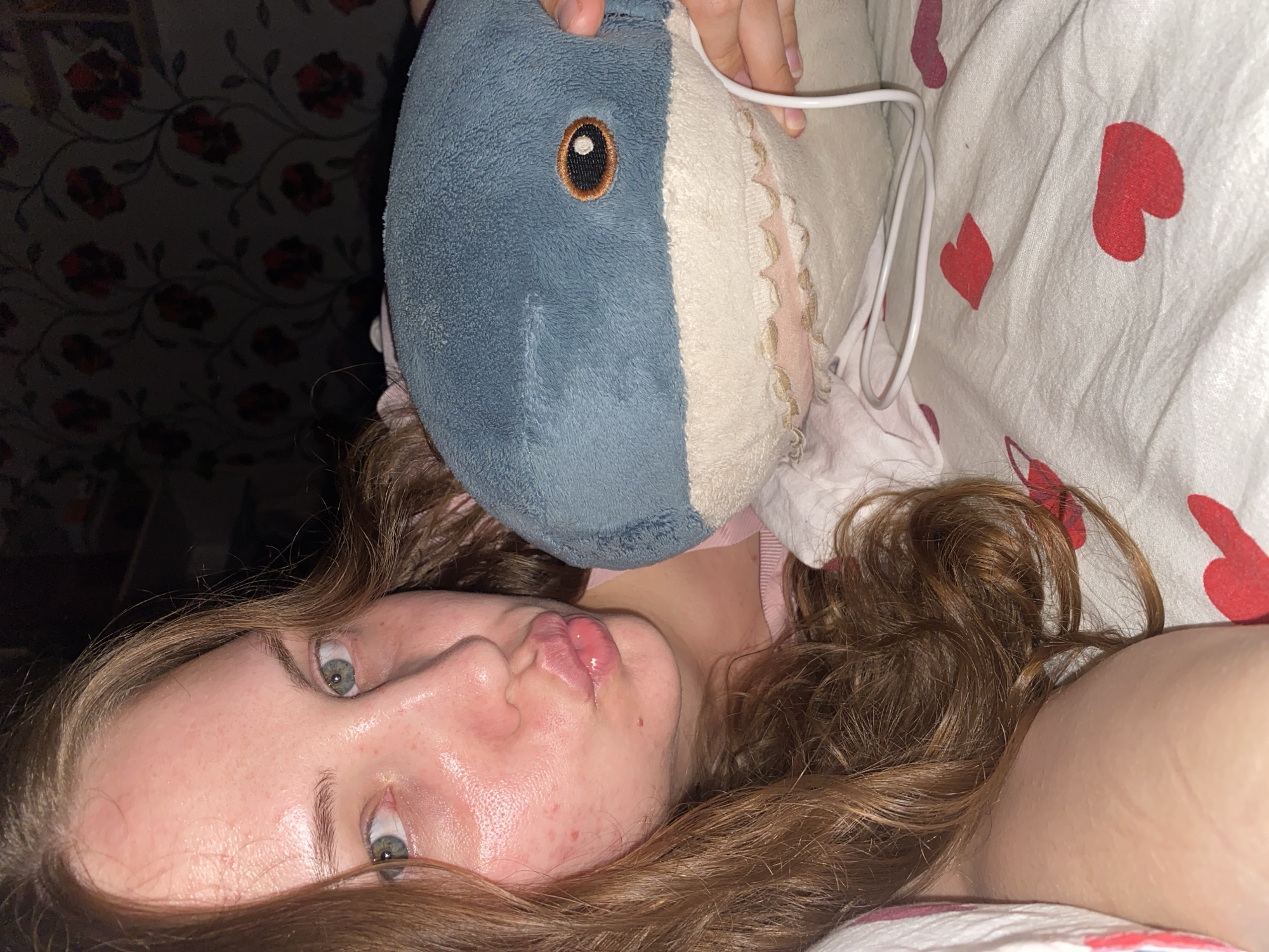 The image shows a close-up of a young woman holding a stuffed shark while lying in bed. The woman has light skin with some redness and small blemishes on her cheeks, and her lips are pursed. She has long, wavy brown hair. The stuffed shark is blue and white with an embroidered orange and black eye and visible fabric teeth. A white cord is visible near the shark. The background consists of a floral patterned wallpaper and a bed with a white sheet decorated with red hearts. The lighting suggests an indoor setting at night or in low light conditions. The location is not specified in the image or its context.
