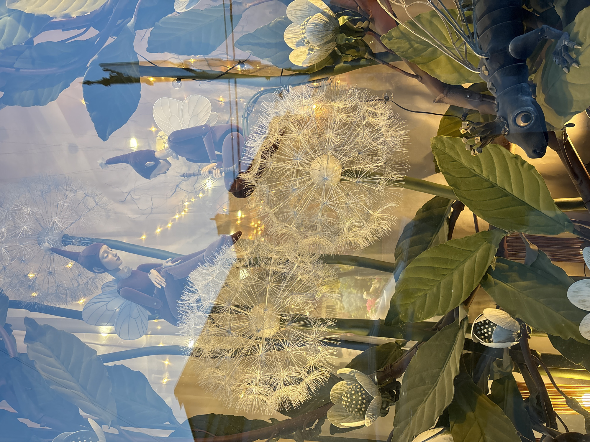 A close-up, high-angle shot depicts a window display in Saint Petersburg, Russia, featuring whimsical fantasy elements. A metal lizard figure hangs upside down amid faux foliage with large, green leaves and white, stylized flowers. A doll with butterfly wings reclines on a large, white dandelion seed head, reflected in the glass. Fairy lights are scattered throughout the display. A second doll is visible in the reflection.
