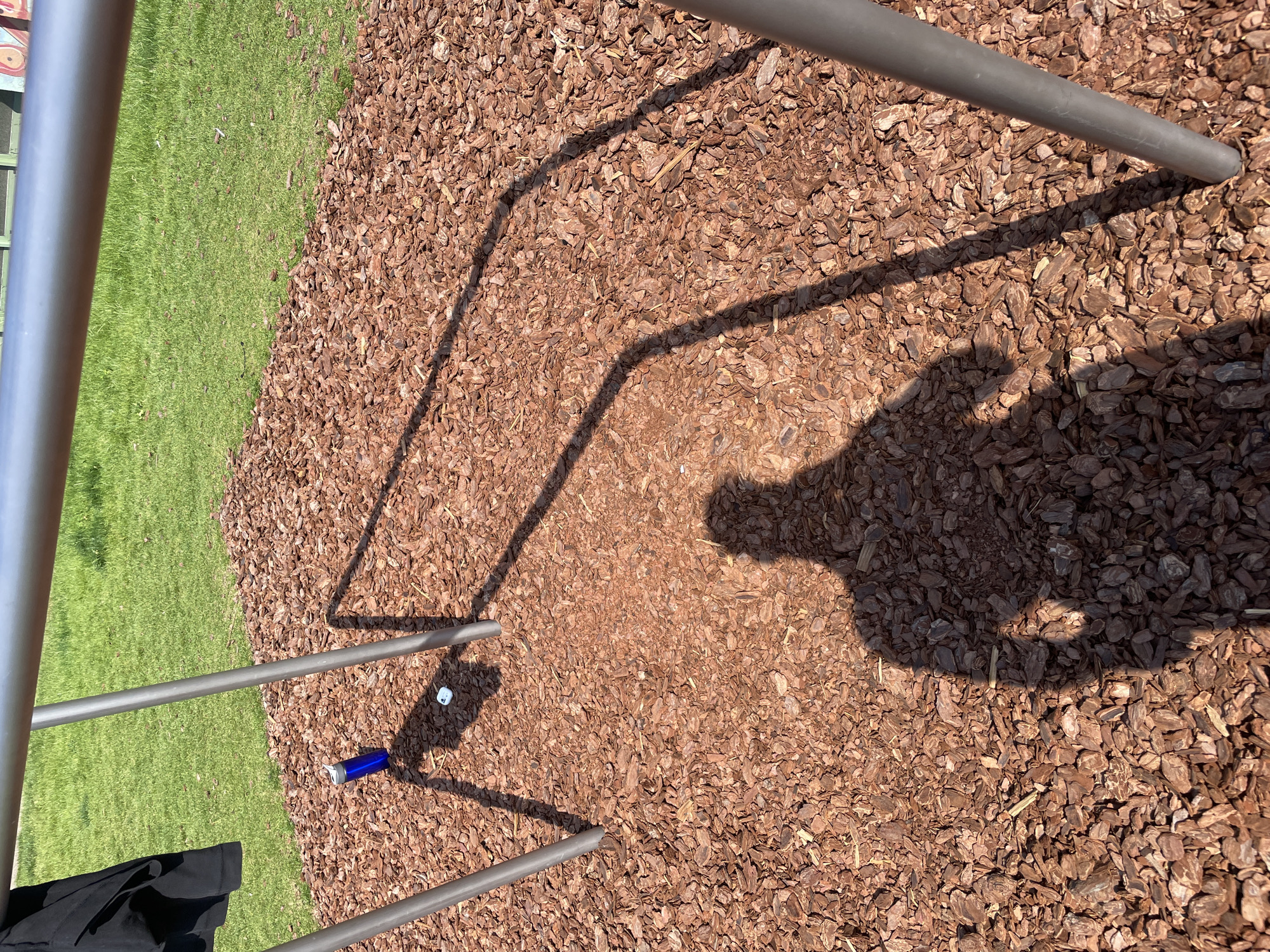 The image depicts a section of ground covered in brown wood chips. Four metal poles, part of a structure, are visible, casting shadows on the wood chips. A patch of green grass is adjacent to the wood chip area on the left side of the frame. A small blue water bottle and a white plastic cap are lying on the wood chips between the poles. The shadow of a person is also visible. The location is not identifiable from the image.