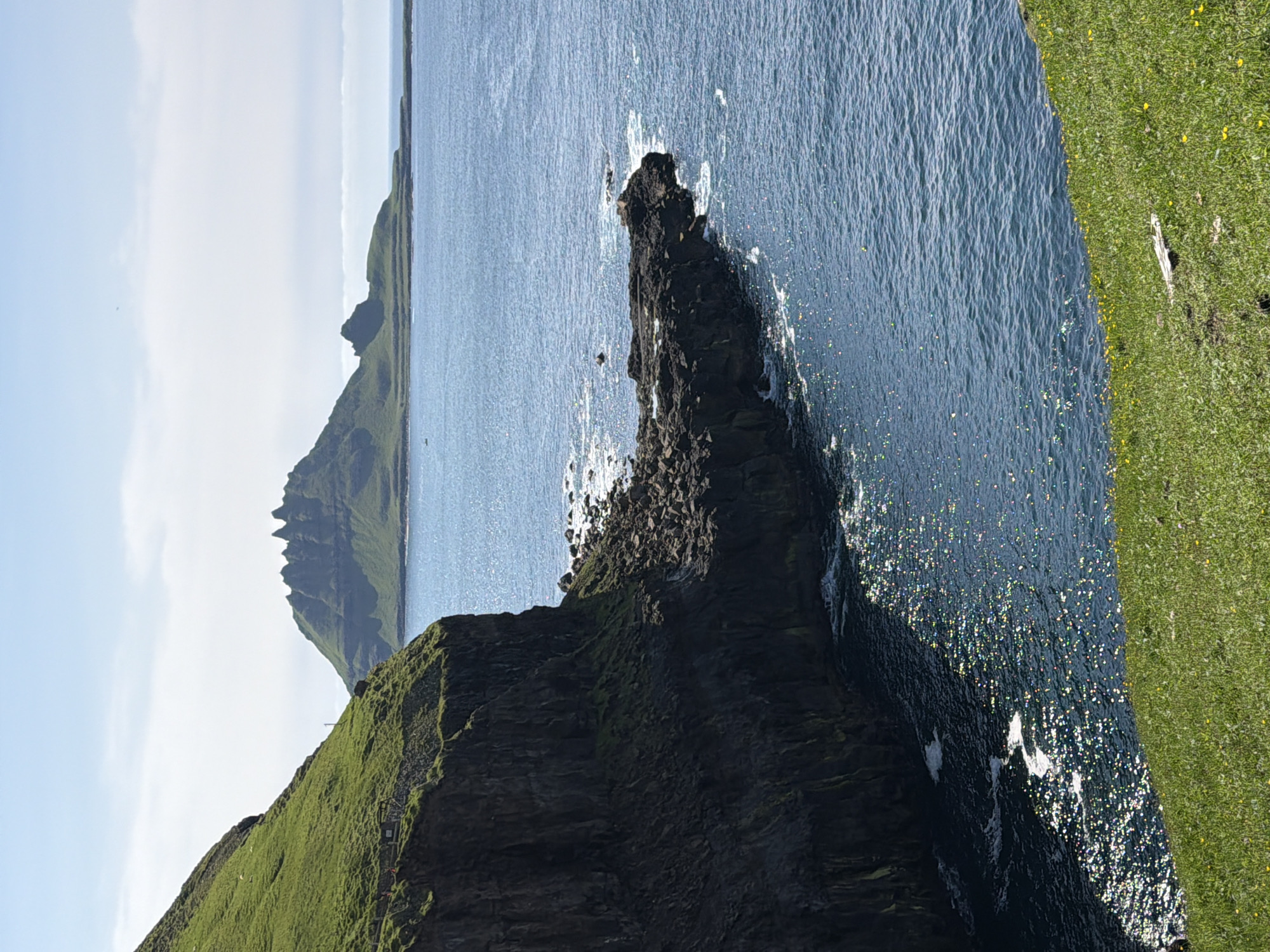 The media file shows a landscape view of the ocean and cliffs in Gásadalur, Faroe Islands. The scene is dominated by rugged, grassy cliffs plunging into a deep blue, rippling ocean. In the foreground, a dark, jagged rock formation juts out into the sea, with waves crashing against its base, creating white foam and glittering highlights on the water's surface. The cliffs are covered in vibrant green grass, dotted with small yellow flowers and some darker patches of rock. In the distance, more imposing, serrated mountains rise from the sea, partially shrouded in soft, hazy light under a pale blue sky with scattered white clouds. The overall impression is one of wild, natural beauty and a sense of vastness. The sun appears to be shining, creating bright reflections on the water, suggesting it is likely daytime. There is no text visible in the image.
