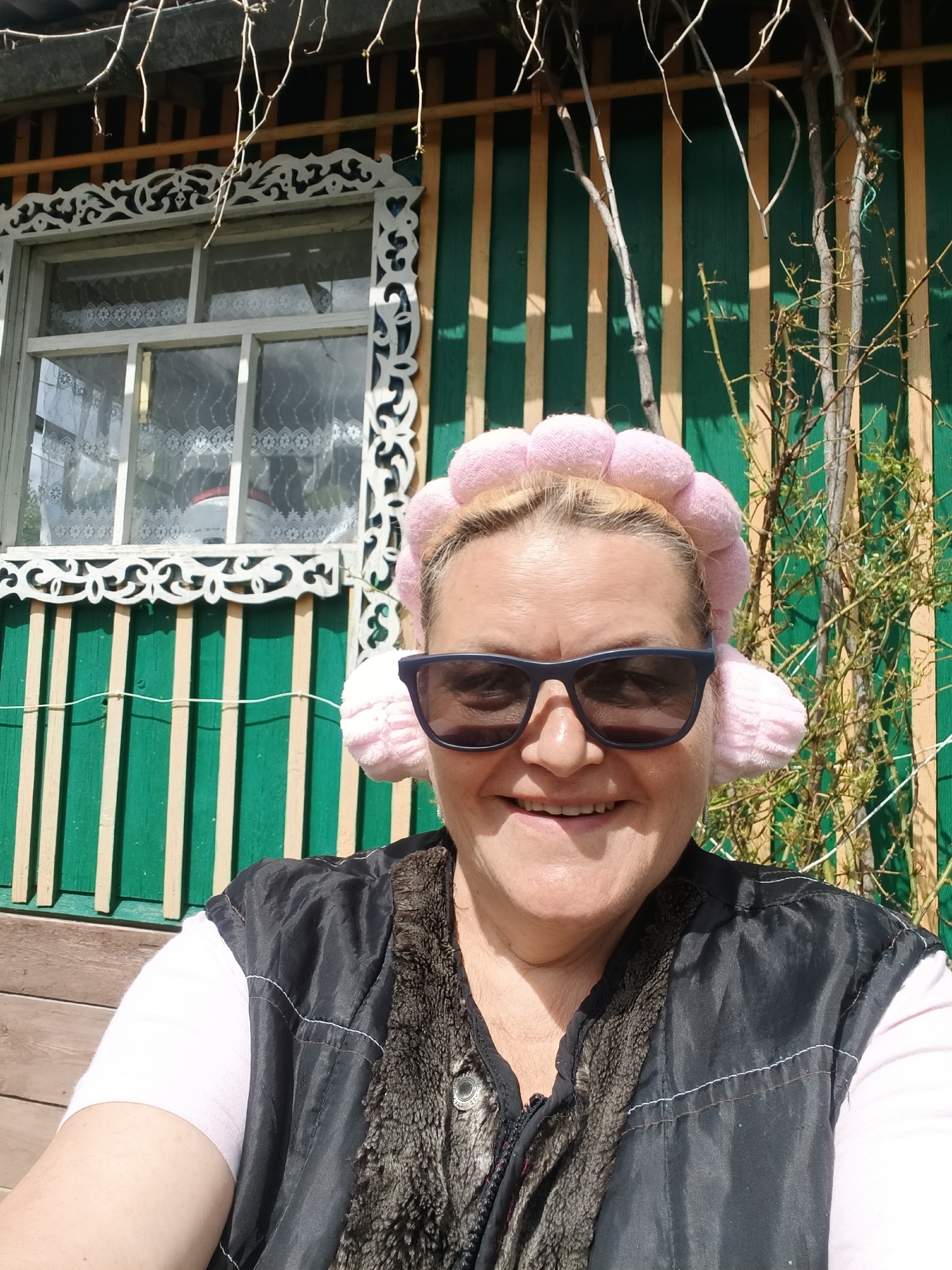 The image shows a woman in sunglasses and a pink pompom headband smiling at the camera. She wears a light pink shirt and a black vest with faux fur lining. She is sitting outdoors in front of a green and tan painted building. The building has a window with elaborate white trim. A small tree or shrub grows near the building. The location is Красный Маяк, Russia.