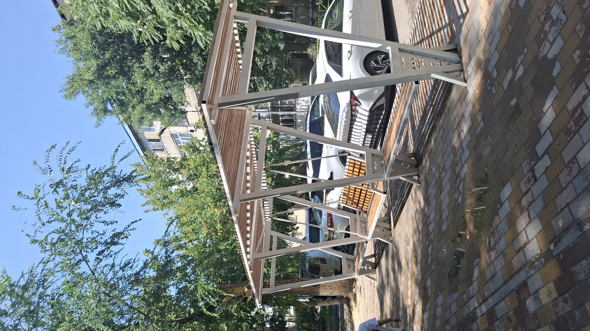 This image depicts an outdoor seating area with a modern, minimalist design, featuring a slatted roof supported by white metal A-frame structures. Underneath the canopy, there are several benches with wooden seats and backrests, some appearing to be swing seats. The seating area is situated on a patterned brick-paved walkway, which shows some standing water and reflections, suggesting recent rainfall. In the background, several cars are parked, primarily white sedans and SUVs. Behind the parked cars, a multi-story building with balconies is visible, partially obscured by lush green trees. The scene is bathed in bright sunlight, casting distinct shadows from the canopy and trees onto the ground. The overall environment appears to be a public space within a city, likely a street or a park area. The presence of the building and the cars suggests an urban setting. The style of the building and the type of vehicles are consistent with a modern urban landscape. Based on the visual cues, this scene is located in the city of Shymkent, Kazakhstan.