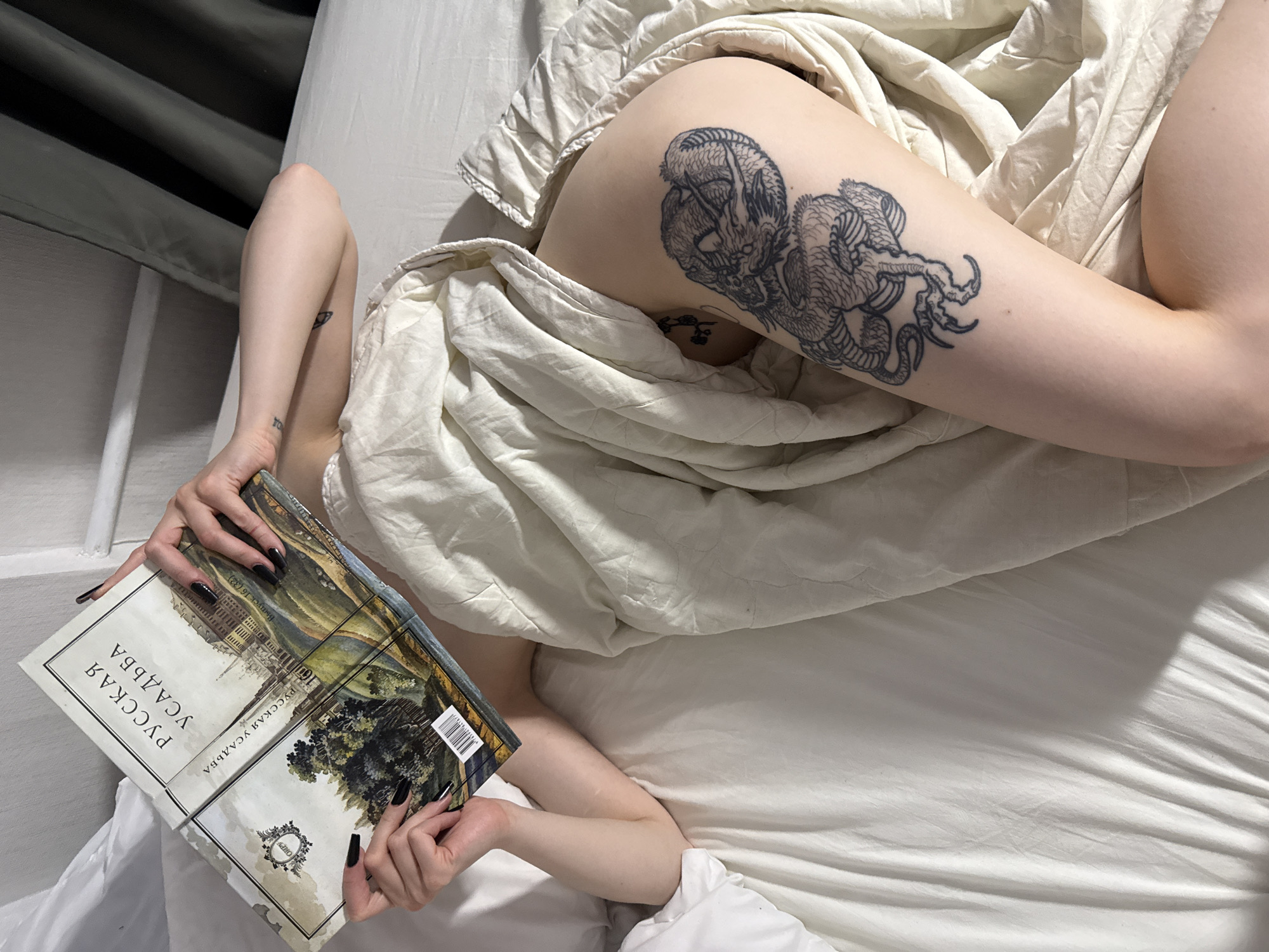 An overhead view captures an individual lying supine on a bed in what is identified as Vniissok, Russia. The person, gender appearing female, is partially covered by a white, textured duvet or blanket. Their head and upper torso are obscured by an open hardcover book titled "РУССКАЯ УСАДЬБА" (Russian Estate) in Cyrillic script, displaying an illustration of a landscape with a large building and trees. Both hands, adorned with dark nail polish, grasp the book. The left forearm bears a small, indistinct tattoo, while the right upper arm shows a faint eye-like tattoo. The individual's right leg, exposed from mid-thigh, features a prominent, detailed black-and-white tattoo of a dragon or mythical creature on the outer thigh, and a smaller, faded floral tattoo higher on the inner thigh. The bed is made with plain white sheets and a white pillow is visible on the left. The background includes a white wall or headboard on the left and dark curtains or a darker wall on the right. Soft, indirect light illuminates the scene, casting subtle shadows on the bedding.
