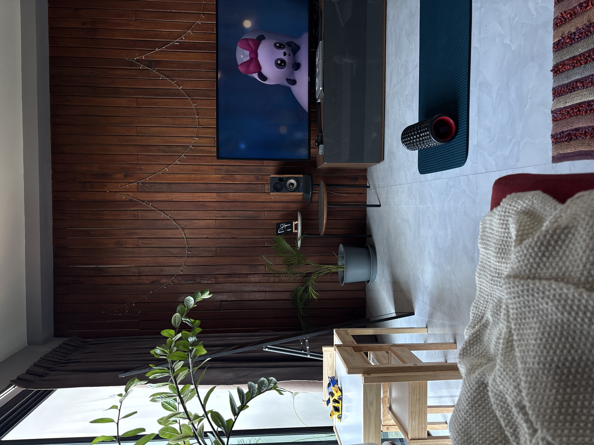 This image captures a bright, modern indoor space, likely a living room or studio in Choeng Thale, Thailand. The central feature is a striking dark brown wooden slatted accent wall, adorned with a crescent-shaped string of delicate fairy lights. A flat-screen television is mounted on this wall, displaying a cartoon image of a light purple panda with a red bow. Below the TV, a dark gray media console supports a single audio speaker placed on a small, round multi-tiered table.
The floor throughout the area is covered with light-toned rectangular tiles. On the right side, a dark teal yoga mat is laid out, holding a black, textured foam roller, suggesting an active area for exercise or stretching. Next to this is a colorful, striped woven rug. A red sofa or armchair, partially visible, is draped with a cream-colored knitted blanket, indicating a comfortable seating zone.
Natural light enters from a large window on the far left, behind dark curtains. In the immediate foreground, a lush, leafy green plant adds to the natural aesthetic. Further into the room, a potted palm-like plant sits on a stand near the wooden wall. A light-colored wooden shelf unit on the right holds children's toys, including a yellow and blue car. A black floor lamp is also partially visible. A small white sign near the potted plant displays two Korean characters, '아' and 'ㄴ'. The scene conveys a calm, functional, and inviting environment, typical of a daytime setting.