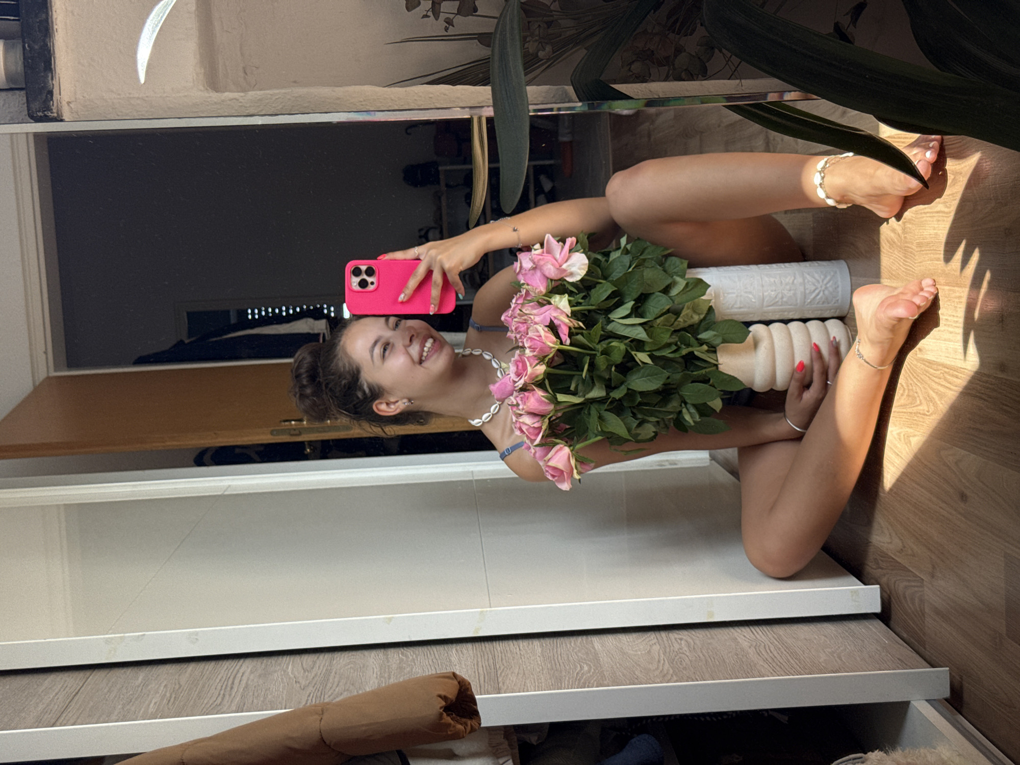 A young woman with brown hair in a bun is sitting on a wooden floor, holding a bouquet of pink roses. She is taking a selfie in a mirror with her phone, which is in a bright pink case. She is wearing a necklace and anklets. Her fingernails are painted pink. The background shows a doorway and a closet. The location is in Bruchköbel, Germany.
