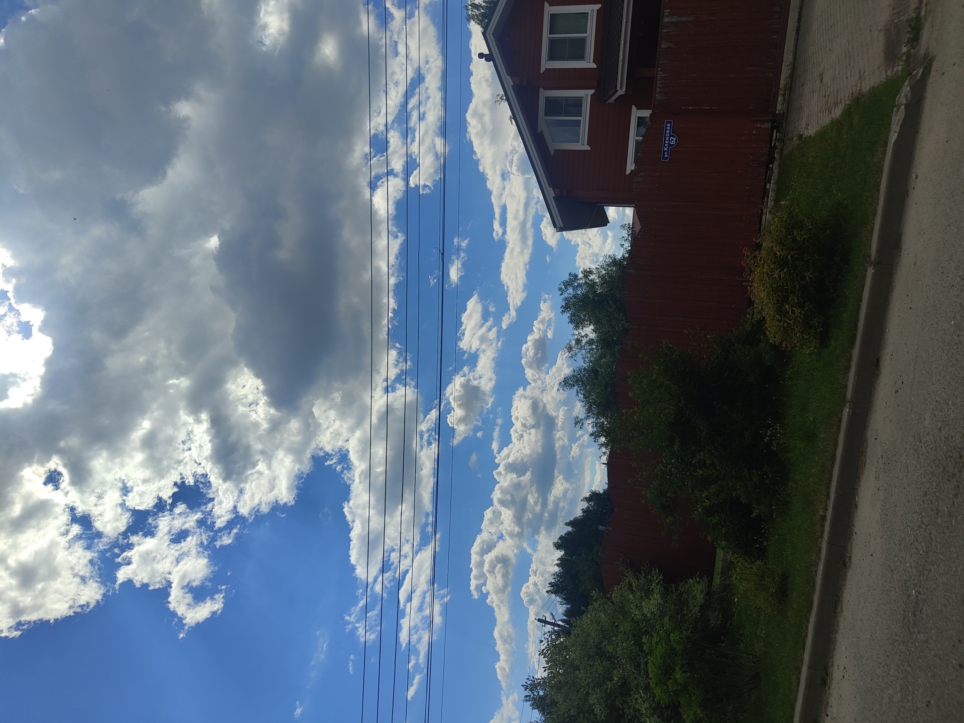 The image captures a view of a street in Tsibino, Russia, on a cloudy day. A two-story, dark red wooden house is visible on the right side of the frame, with white-trimmed windows. A tall, dark red wooden fence runs along the property line. The sky above is a mix of bright blue and heavy grey clouds, with several power lines stretched across it. Dense green foliage and trees are present on the left side of the image, bordering the fence. A paved driveway leads to the house, and a strip of grass and curb is visible in the foreground. A street sign is affixed to the fence, reading "ул. Кленовая 62". No people are present in the scene.
