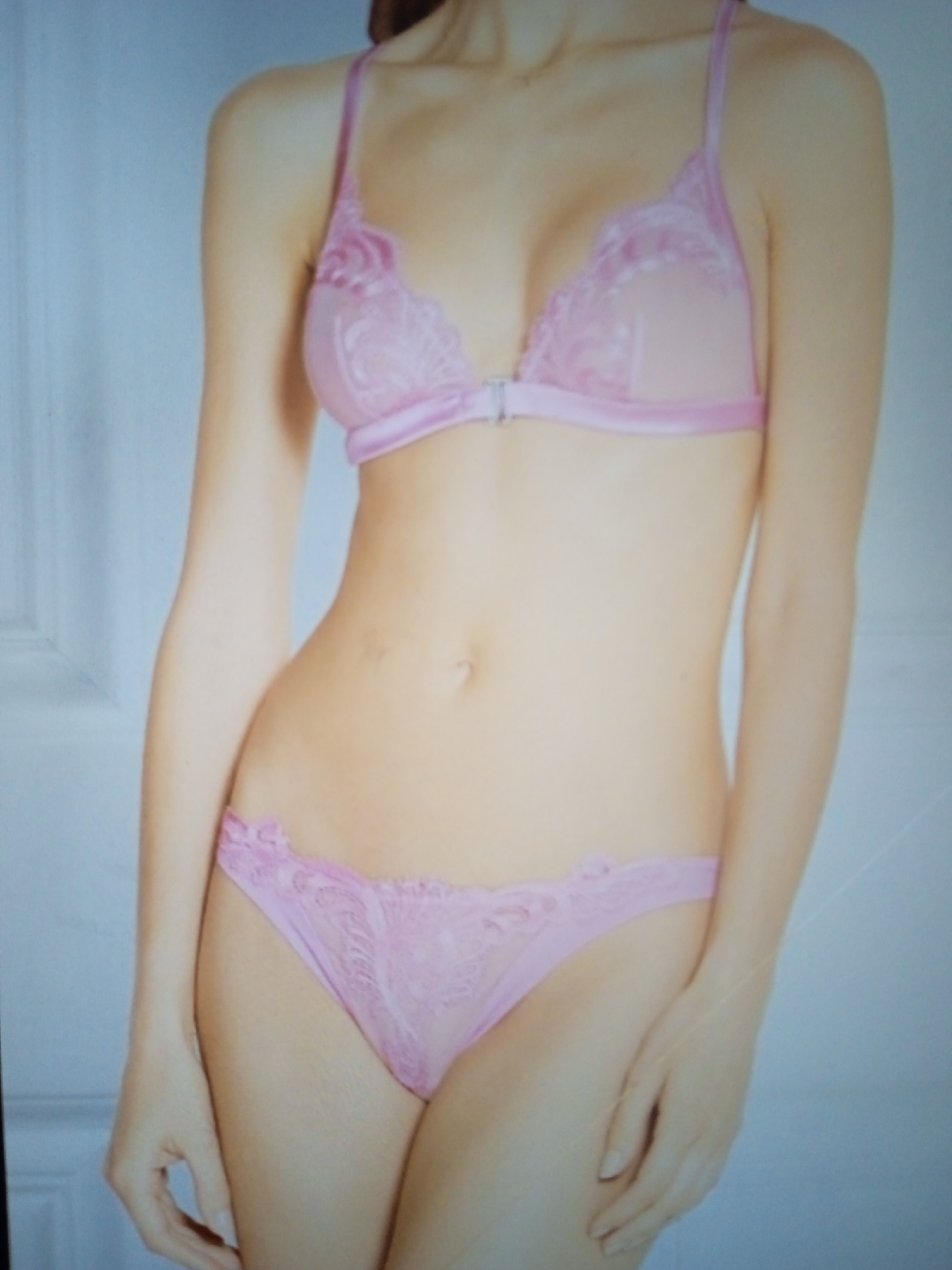 A full-body, medium-shot, eye-level photograph shows a person wearing a pink lace bra and matching pink lace underwear. The person is standing in front of a plain, light blue wall with a subtle vertical line pattern visible on the left side of the image, possibly indicating a door or wall seam. The lighting is soft and even, highlighting the person's skin tone and the delicate details of the lingerie. The person's arms are slightly away from their body, and their hands are resting by their sides. The image is cropped slightly, with the top of the person's head and their feet not visible. There is no discernible context or location within Chernivtsi, Ukraine, as the background is generic and offers no specific identifying features.