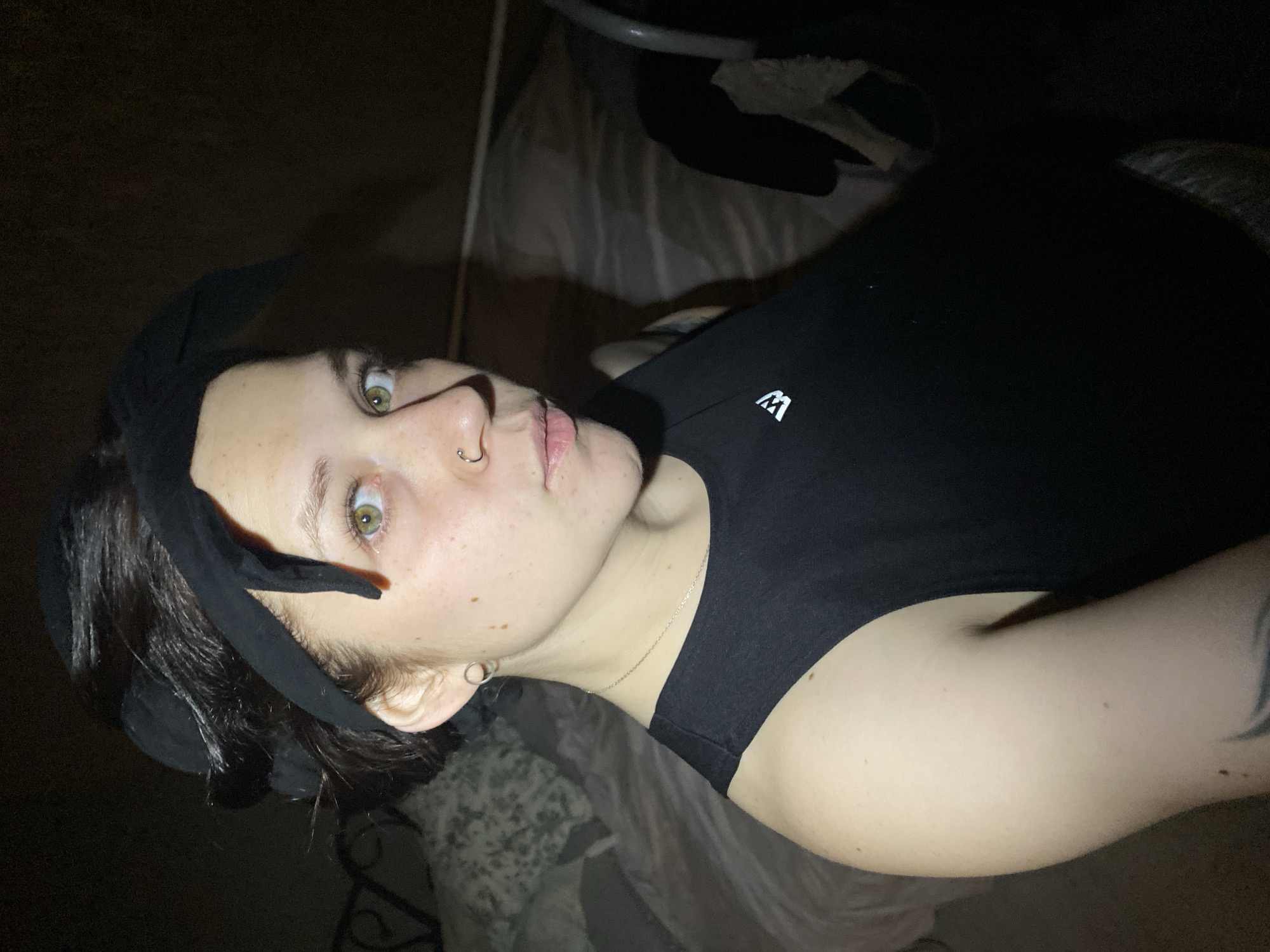 A close-up, low-angle shot of a person wearing a black tank top and a black headband with a bow on top. The person has fair skin, short dark hair, light green eyes, and a piercing in their left nostril. The background appears to be a dark, dimly lit room with a bed in the background. The location is unknown and cannot be verified as Chaikovsky, Russia.