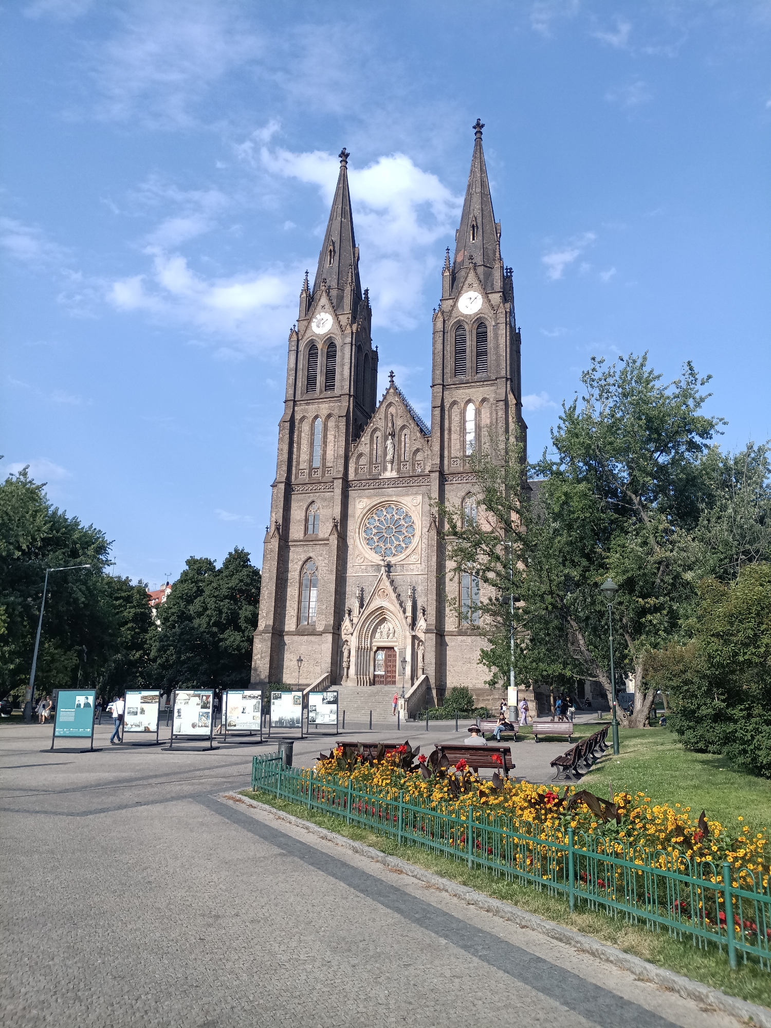 This image depicts the Church of St. Ludmila, located in the Vinohrady district of Prague, Czechia. The church is a prominent Gothic Revival structure with two tall spires, a rose window above the main entrance, and detailed stone carvings. In the foreground, there is a paved walkway and a grassy area bordered by a green metal fence adorned with yellow and red flowers. Several people are visible in the background, some walking on the plaza in front of the church and others sitting on benches in the grassy area. There are also several display boards set up on the plaza. The sky is blue with scattered clouds, suggesting it is daytime. The overall scene captures a sunny day in a public space, highlighting the architectural grandeur of the church and the pleasant urban environment.