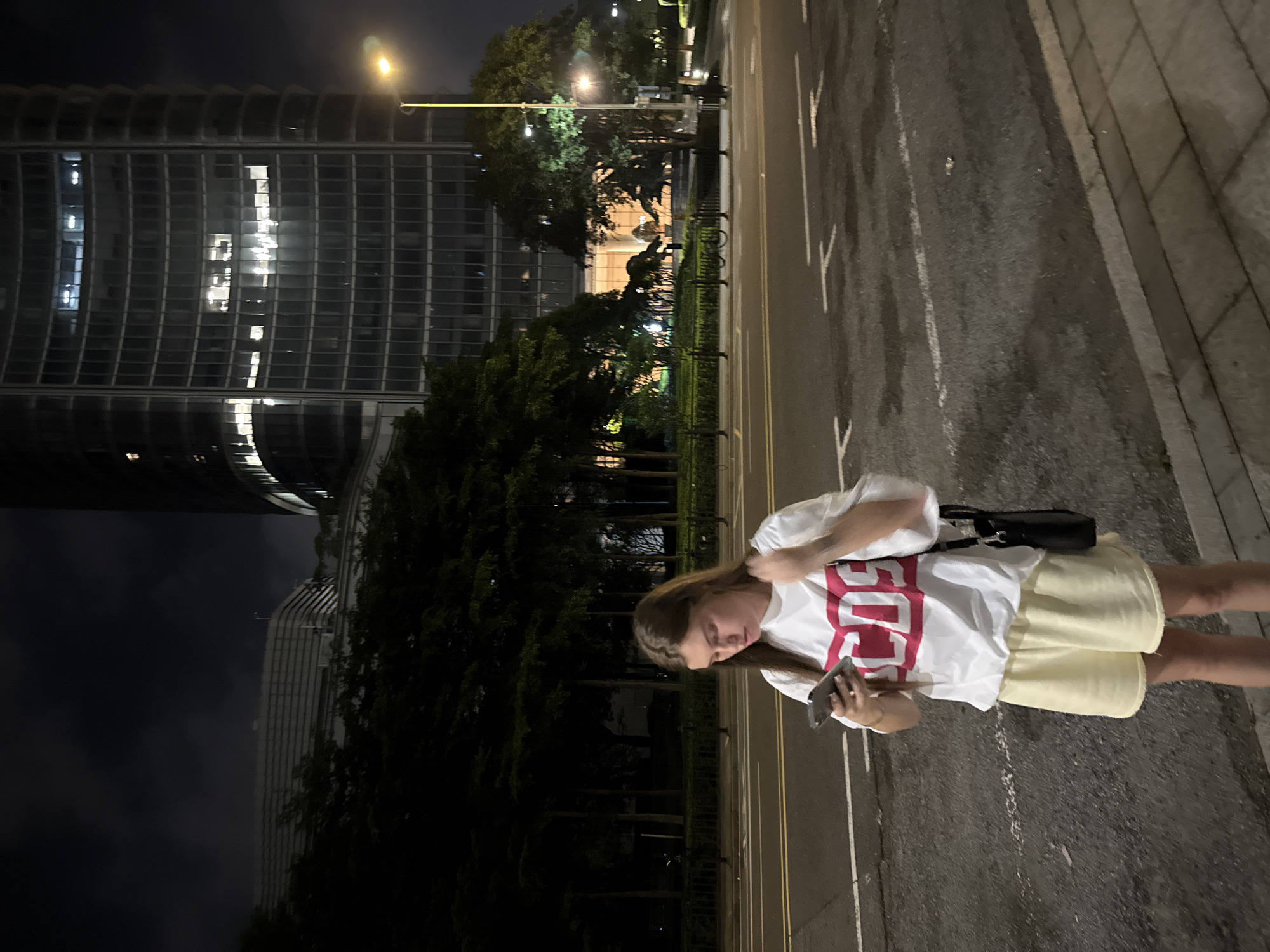 A young woman stands on a street at night, holding a smartphone. She is wearing a white t-shirt with a large red graphic that appears to read "SOS", and light yellow shorts. A black purse is slung over her shoulder. The street is wet, suggesting it may have recently rained, and there are white lane markings on the asphalt. To her left, a dark building with numerous illuminated windows rises into the night sky. Trees line the area between the building and the street. The scene is illuminated by streetlights and the lights from the building. The woman's gaze is directed downwards at her phone, and her expression is neutral. The time appears to be late evening or night, and the weather is likely damp but not actively raining. The location is identified as Haizhu District, China.