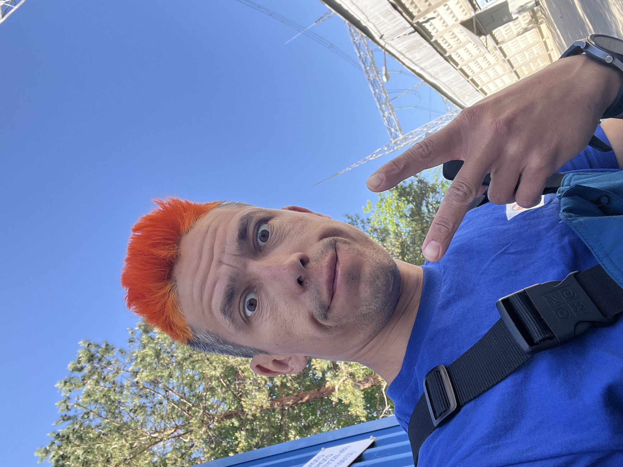 A man with orange hair is taking a selfie outside in Chaikovsky, Russia. He is wearing a blue t-shirt and a black watch. He is pointing upwards with his right hand, and a tree and a metal structure are visible in the background. The sky is blue and clear.