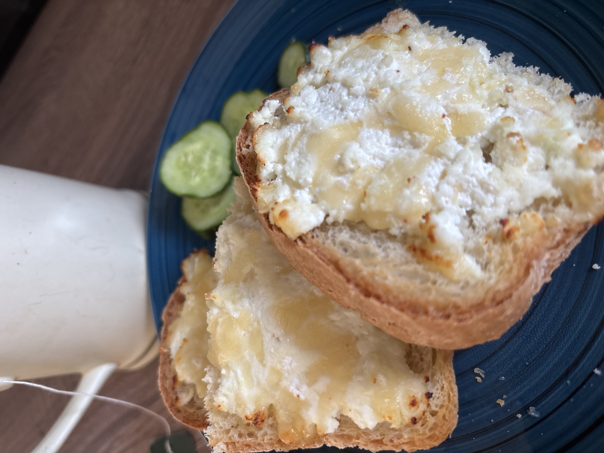 A close-up, eye-level shot showcases a blue plate of toasted bread slices topped with a creamy white spread and drizzled with honey. The bread appears rustic, with a golden-brown crust. Several round, green cucumber slices are arranged on the side of the plate. In the background, out of focus, is a cream-colored mug with a tea bag string hanging out, placed on a wooden surface. The scene suggests a casual breakfast or snack. There is no information or visual cues in the image to suggest a specific location within Naberezhnye Chelny, Russia.