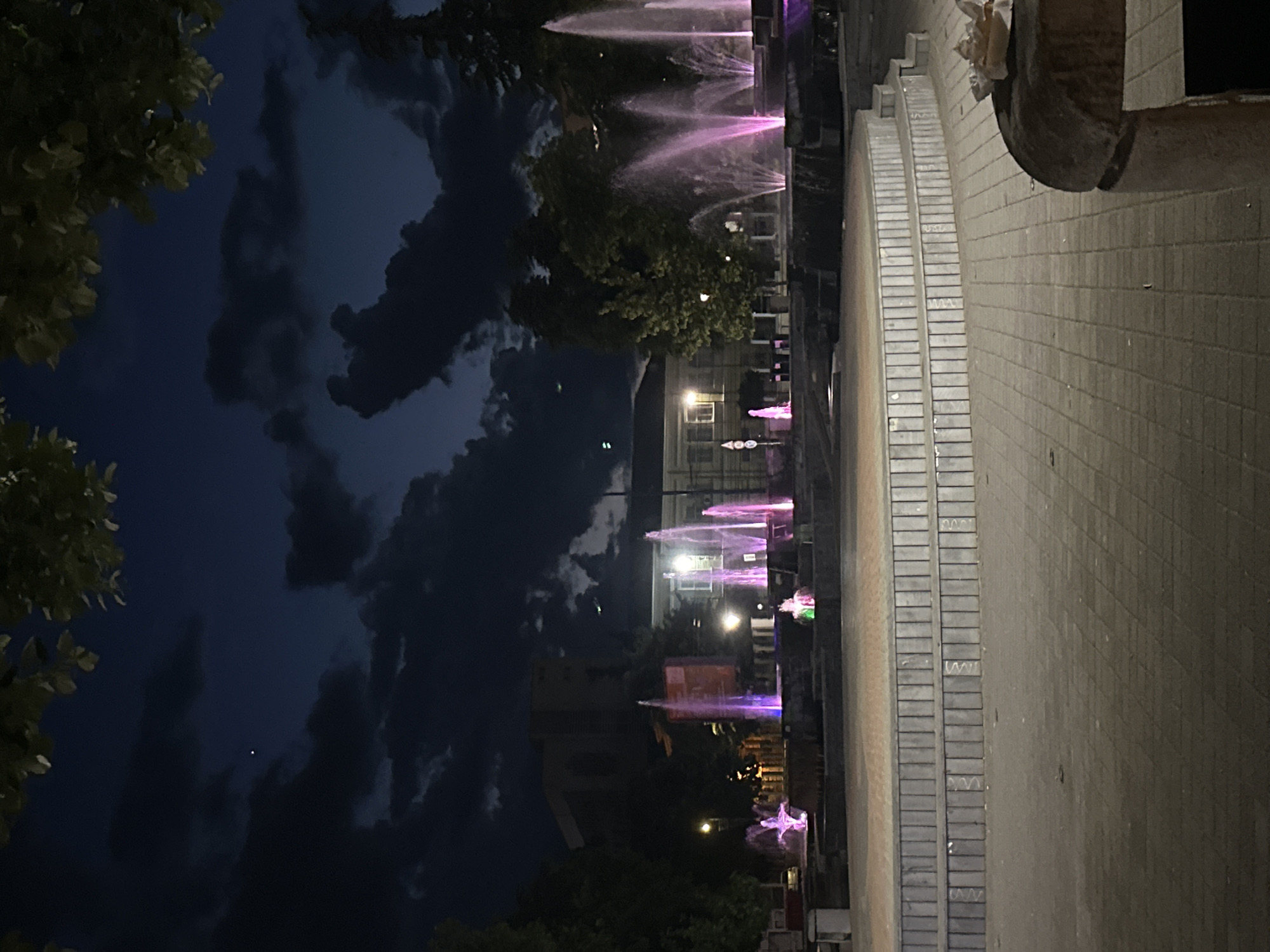The image shows a row of illuminated fountains in Pazardzhik, Bulgaria. The fountains are arranged in a line, and the water is lit up with purple and pink lights. Trees and clouds are visible in the background. A paved area is on the right side of the image, leading to a building. The scene is at night, and the sky is dark with clouds.