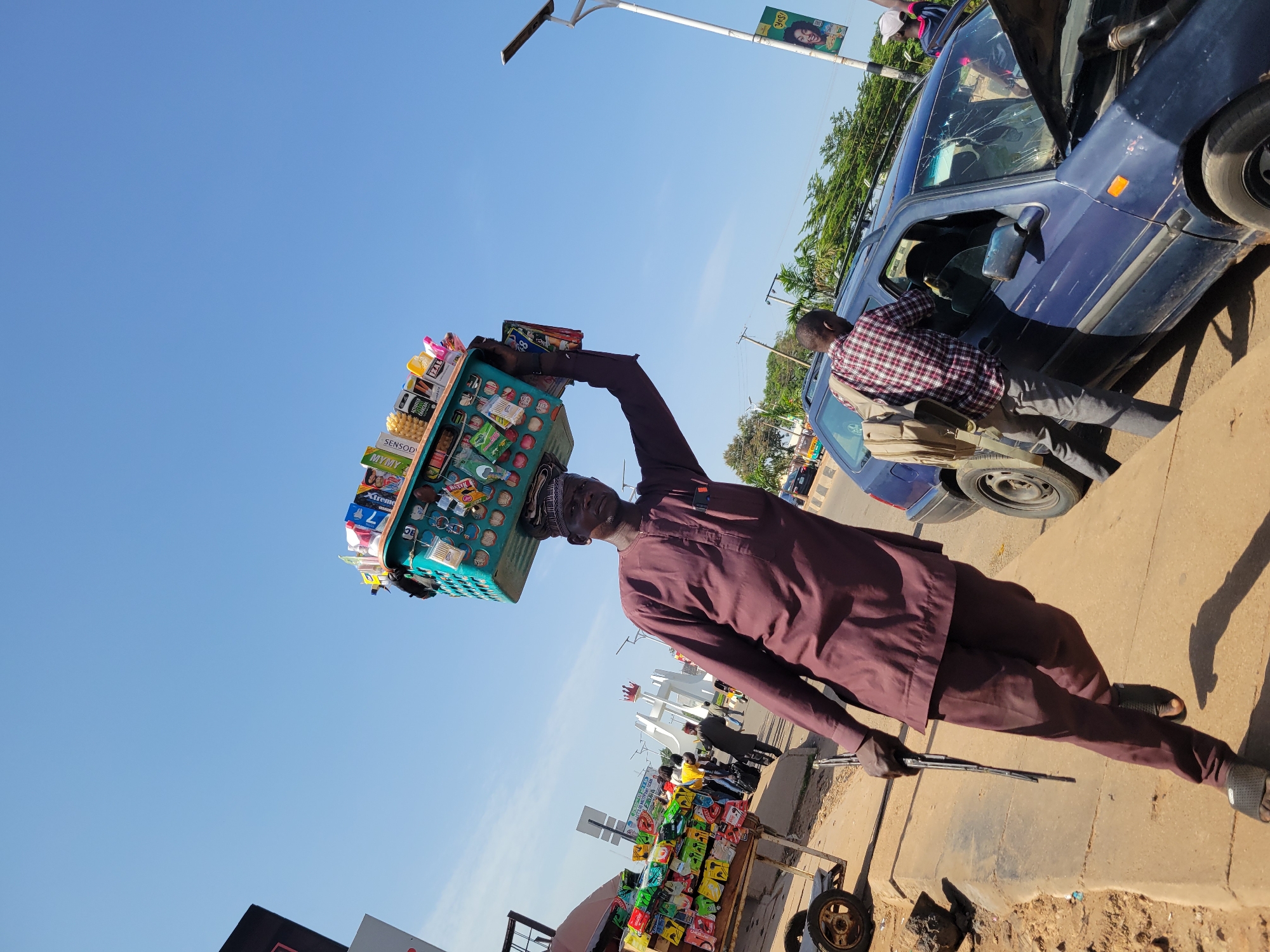 The image depicts a street scene in Jos, Nigeria, under a clear blue sky. A man, dressed in a maroon traditional outfit and a cap, walks along a sidewalk carrying a teal basket filled with various packaged goods on his head. He holds a long, flat object, possibly a windshield wiper, in his right hand.
To the left of the man, a mobile stall displays numerous colorful snack packets. In the background, people are visible on the sidewalk, and a white archway structure stands in the distance. A blue car is parked on the right side of the road, its windshield visibly cracked. Another person, wearing a plaid shirt and a backpack, is interacting with the driver of the blue car. Trees and utility poles line the street, with a lamppost featuring a solar panel and a poster on the right. The ground is paved with concrete and dirt. The overall impression is of a busy urban environment during the daytime.