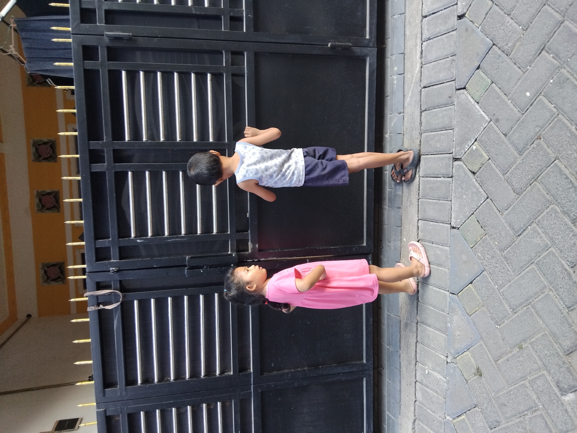 Two young children, a girl and a boy, stand facing a black metal gate with horizontal bars in front of a building. The girl, on the left, wears a pink dress and pink sandals. She has her hands on her hips. The boy, on the right, wears a white tank top with a light blue pattern and dark blue shorts, and dark sandals. He has his hands on the gate, looking through it. The ground is paved with grey interlocking bricks arranged in a herringbone pattern. The scene appears to be in front of a residential building, possibly located in Kepuh Kiriman, Indonesia, given the architectural style and context. The sunlight suggests it is daytime.