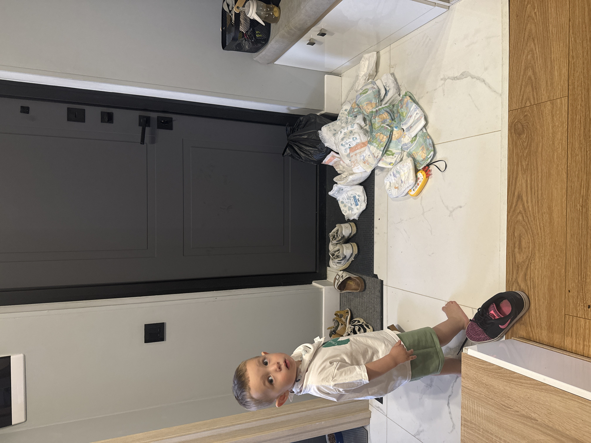 The scene is an indoor shot taken inside a residence. A young child, approximately 2-3 years old, stands in the lower part of the frame, looking towards the viewer with a curious expression. The child wears a white shirt and green shorts.
Behind the child, on the floor, is a gray rug. A collection of used diapers is haphazardly piled on the floor near a black trash bag. A few pairs of shoes are scattered on the rug.
The background includes a gray door, a white wall with an electrical outlet, and a wooden cabinet along the right side of the frame. The flooring appears to be light-colored marble or tile.
There is insufficient information to place the scene in the city of Odintsovo, Russia.