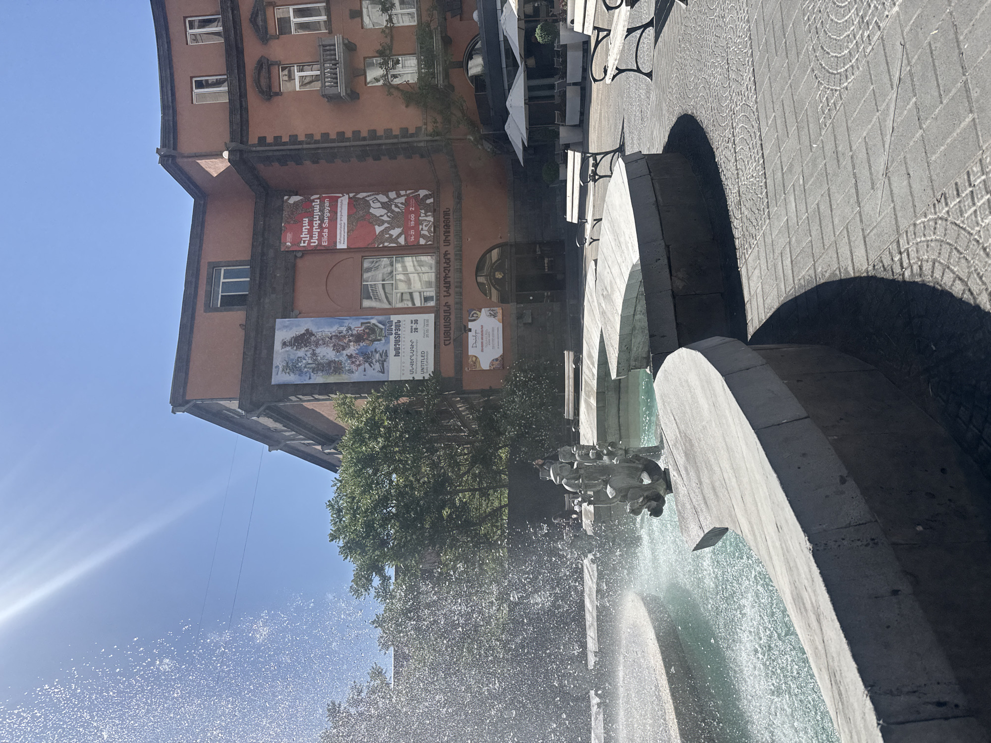 The image shows a fountain in Yerevan, Armenia. The fountain is made of stone and has multiple tiers. Water is spraying out of the fountain, creating a mist in the air. Behind the fountain is a building with several posters on it advertising art exhibitions. The sky is blue and the sun is shining. There are trees visible in the background. Tables and benches are adjacent to the fountain. The image is taken from a slightly low angle, looking up at the fountain and the building.