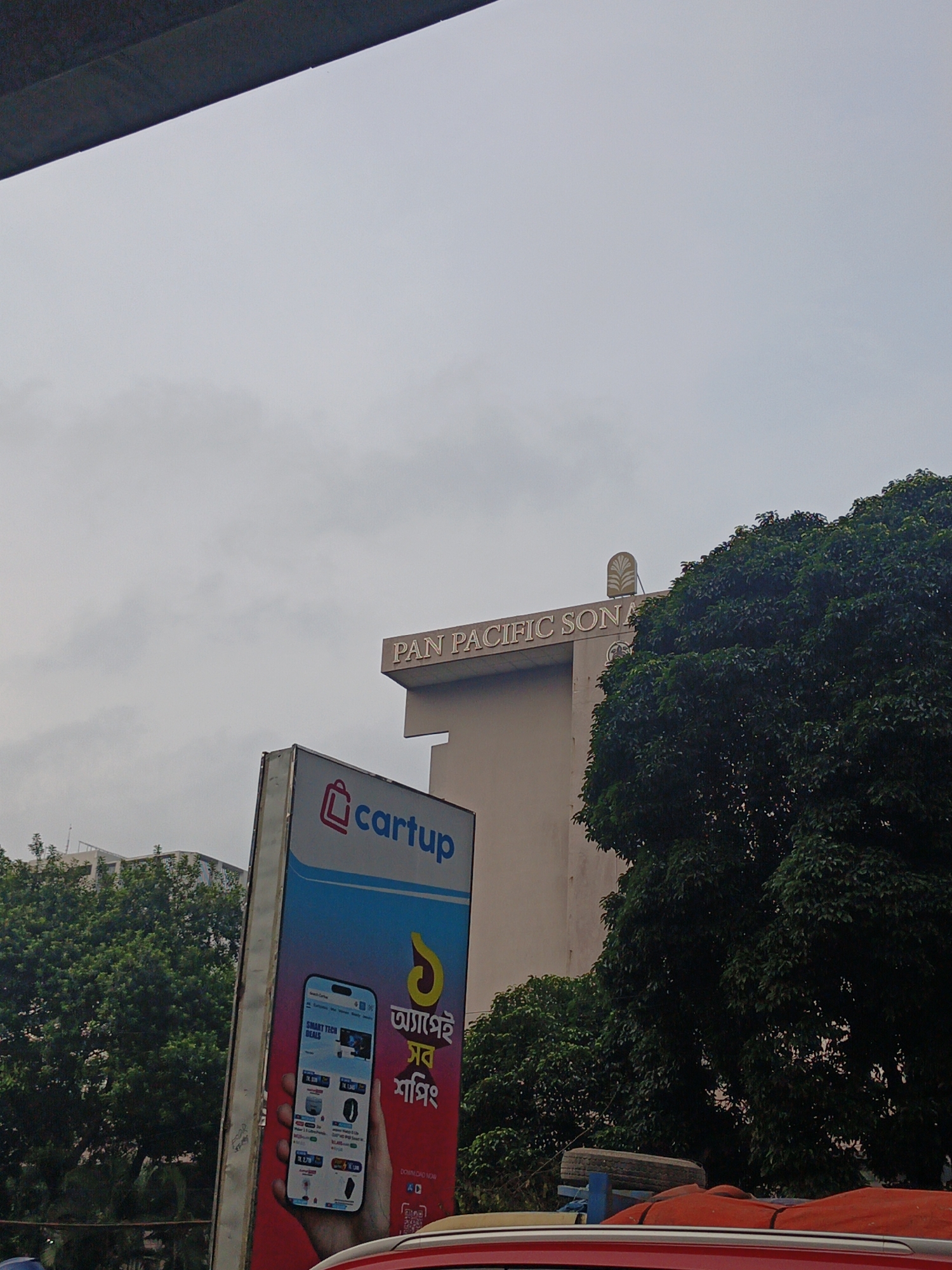 The image shows a street scene in Dhaka, Bangladesh, under a cloudy sky. A prominent billboard for "cartup" is visible on the left, displaying a smartphone with various deals. To the right of the billboard, a building with "PAN PACIFIC SON" inscribed on it is partially obscured by a large, dense green tree. The building appears to be a hotel or office building. In the foreground, a red vehicle is partially visible, with an orange tarp and a spare tire on top of another vehicle behind it. The overall impression is of a busy urban environment.