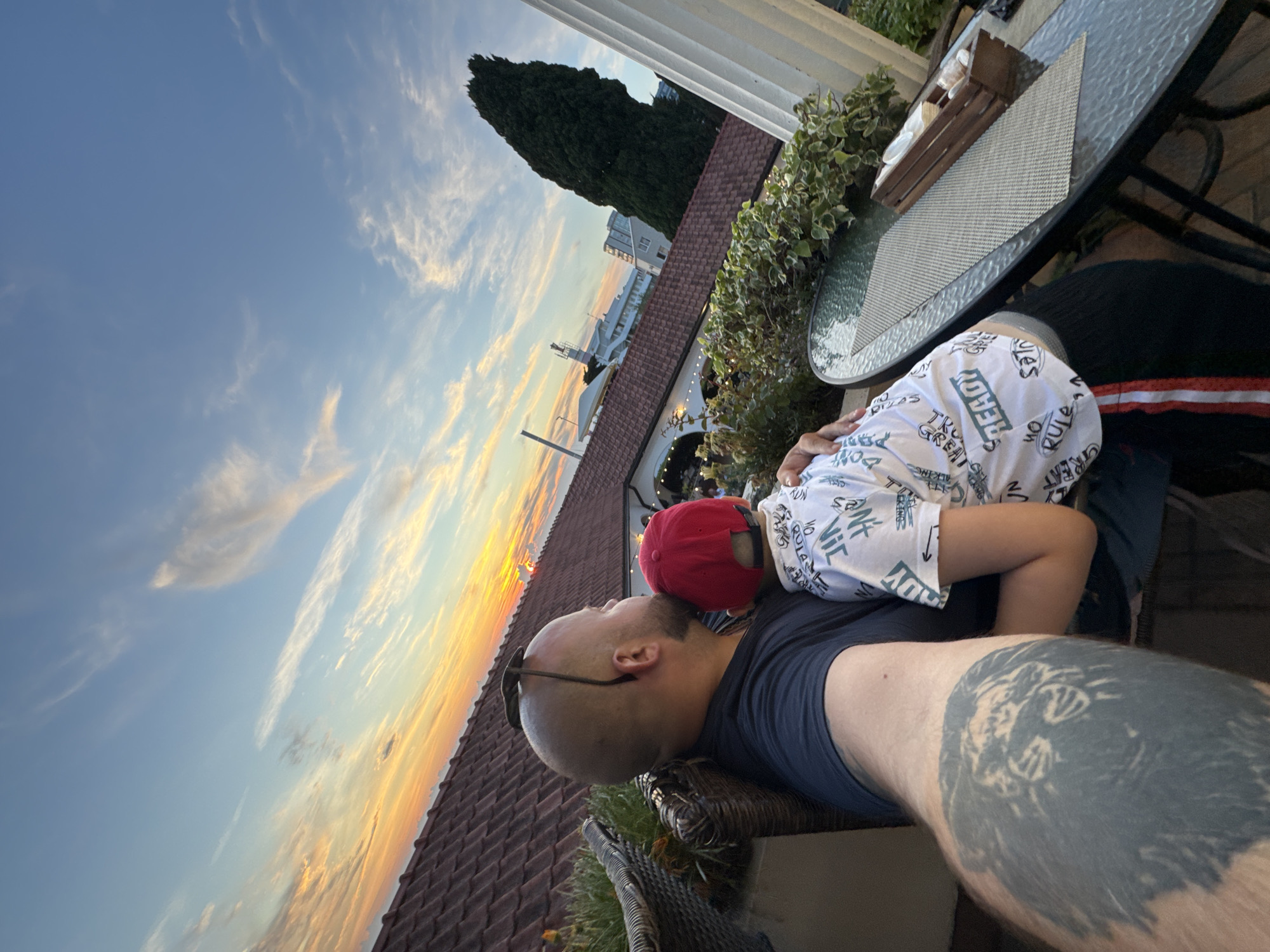 In a snapshot taken from a high vantage point in Sochi, Russia, a man and a child are captured in an embrace against the backdrop of a vibrant sunset. The man, bald and sporting a tattoo of a lion on his arm, rests his head against the child's. The child, wearing a red cap and a white shirt adorned with text, is nestled into the man's arms.
The scene unfolds on a terrace, suggested by the presence of a glass-topped table with a placemat and a wooden tray holding napkins and cutlery. Lush green plants frame the terrace, with a tall, dark green coniferous tree standing sentinel in the background. Beyond the foliage, a row of buildings with terracotta roofs is visible, hinting at a coastal city setting. The sky above is a spectacle of color, with hues of orange, yellow, and blue blending as the sun dips below the horizon. wispy clouds add texture to the sky, illuminated by the fading sunlight. In the distance, a white structure, possibly a lighthouse or a tower, stands out against the twilight sky, and a large ship or boat is anchored offshore. The overall atmosphere is one of tranquility and connection, highlighted by the warm glow of the sunset.