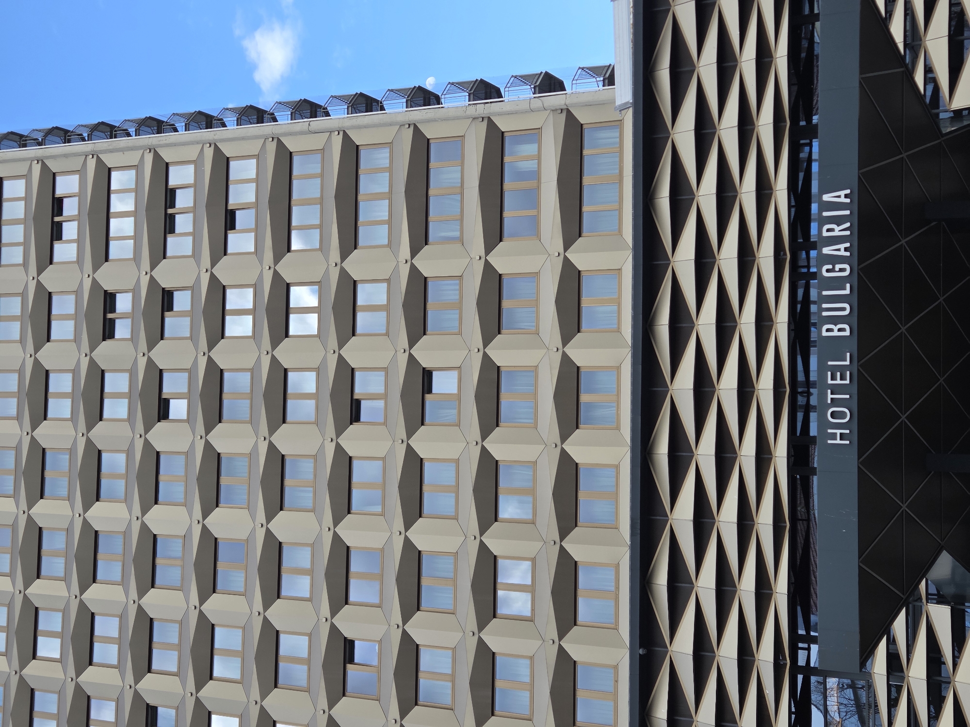 The image displays a detailed view of the Hotel Bulgaria, situated in Burgas, Bulgaria. The upper portion of the building presents a multi-story facade characterized by a repeating pattern of rectangular windows. Each window is recessed within a geometric frame, creating a tessellated appearance with light beige panels forming the primary surface, accentuated by darker brown or grey elements around the window openings. The windows themselves possess light brown frames and reflect the clear blue sky, which contains scattered white cumulus clouds. On the right side of the tower, a vertical stack of individual balconies with dark metal railings and glass panels is visible. One window on the sixth floor from the bottom, third from the left, is partially open.
Below the main tower, a distinct lower section features an elaborate geometric facade composed of numerous interlocking triangular or diamond-shaped panels in alternating light and dark hues. A prominent horizontal sign, dark grey in color, spans this section, displaying the white uppercase text "HOTEL BULGARIA." Directly beneath the sign, a dark, geometrically patterned canopy extends outward. Reflections in the glass surfaces of the lower section show distorted images, including tree branches. The scene is illuminated by natural daylight. No individuals are visible, and no actions or interactions are depicted within the frame.