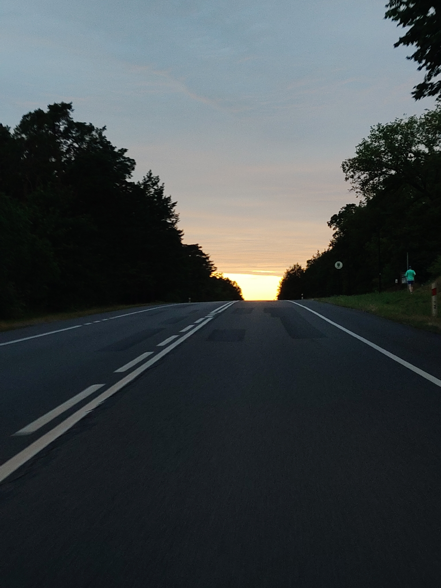 The image depicts a road with white markings ascending towards a bright horizon, likely at sunset or sunrise. The road is flanked by dark, dense trees on both sides. On the right side of the road, a person in a green shirt and dark shorts is visible walking away from the camera. There are traffic signs and posts along the roadside. The sky is a soft gradient of blue and orange, with faint clouds. The overall impression is of a rural or semi-rural setting, possibly in or near Rajszew, Poland, given the context provided, during twilight.