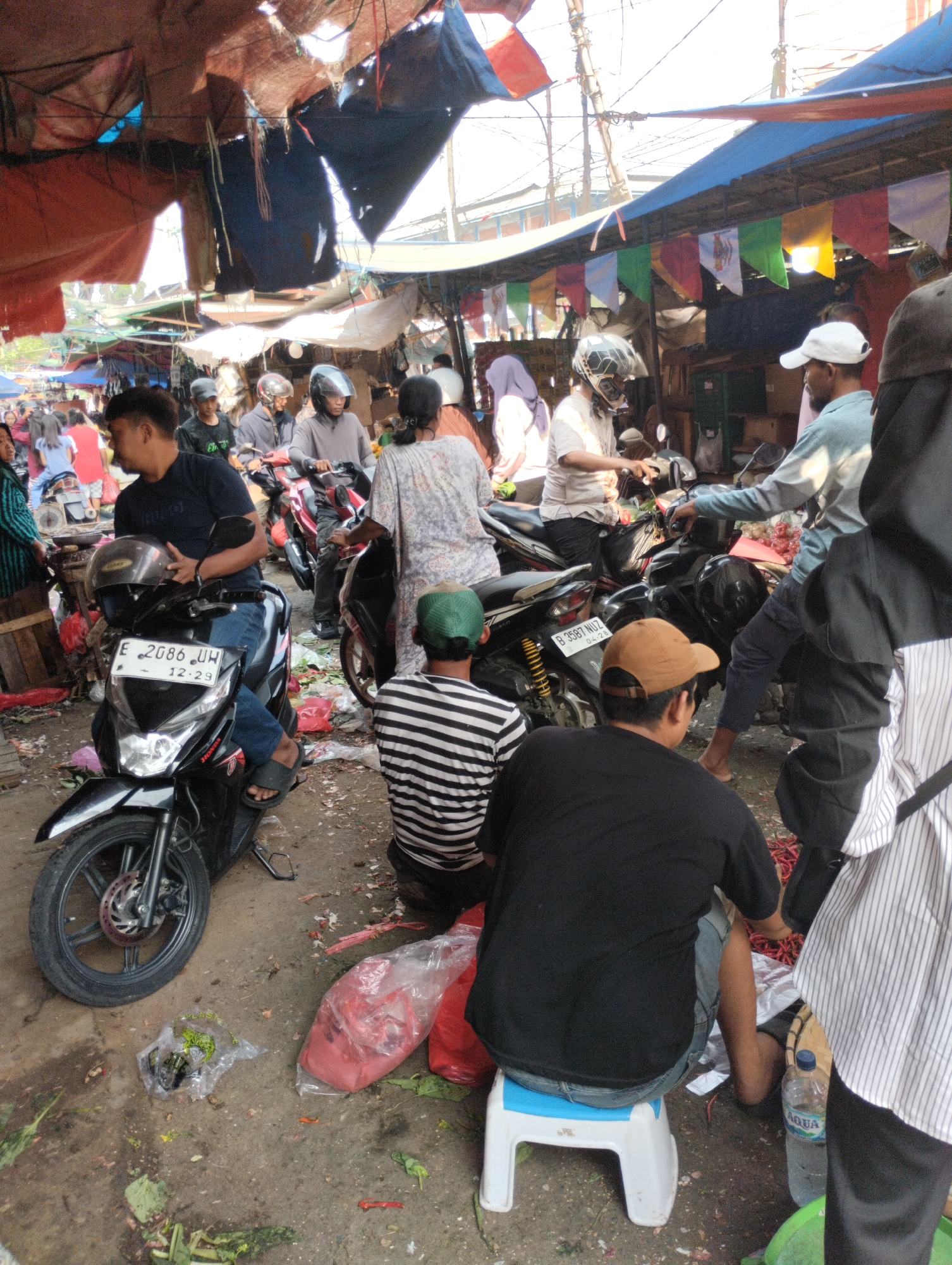A busy street market in Bekasi, Indonesia, is depicted. The scene is crowded with people and several motorcycles. Tents and tarps, some tattered, provide shade overhead, creating a cluttered and somewhat disorganized environment. Vendors appear to be selling goods from stalls. In the foreground, a man in a black t-shirt and jeans sits on a small white stool. To his right, another man in a black t-shirt and blue jeans is seated on a black motorcycle, holding his helmet. Behind him, a motorcycle has a license plate that reads "E 2086 UH". Other individuals are seen on motorcycles and walking through the market. The ground is covered in debris, including plastic bags and fallen produce. Colorful flags are strung between some of the market stalls. The overall impression is one of a vibrant, bustling marketplace.