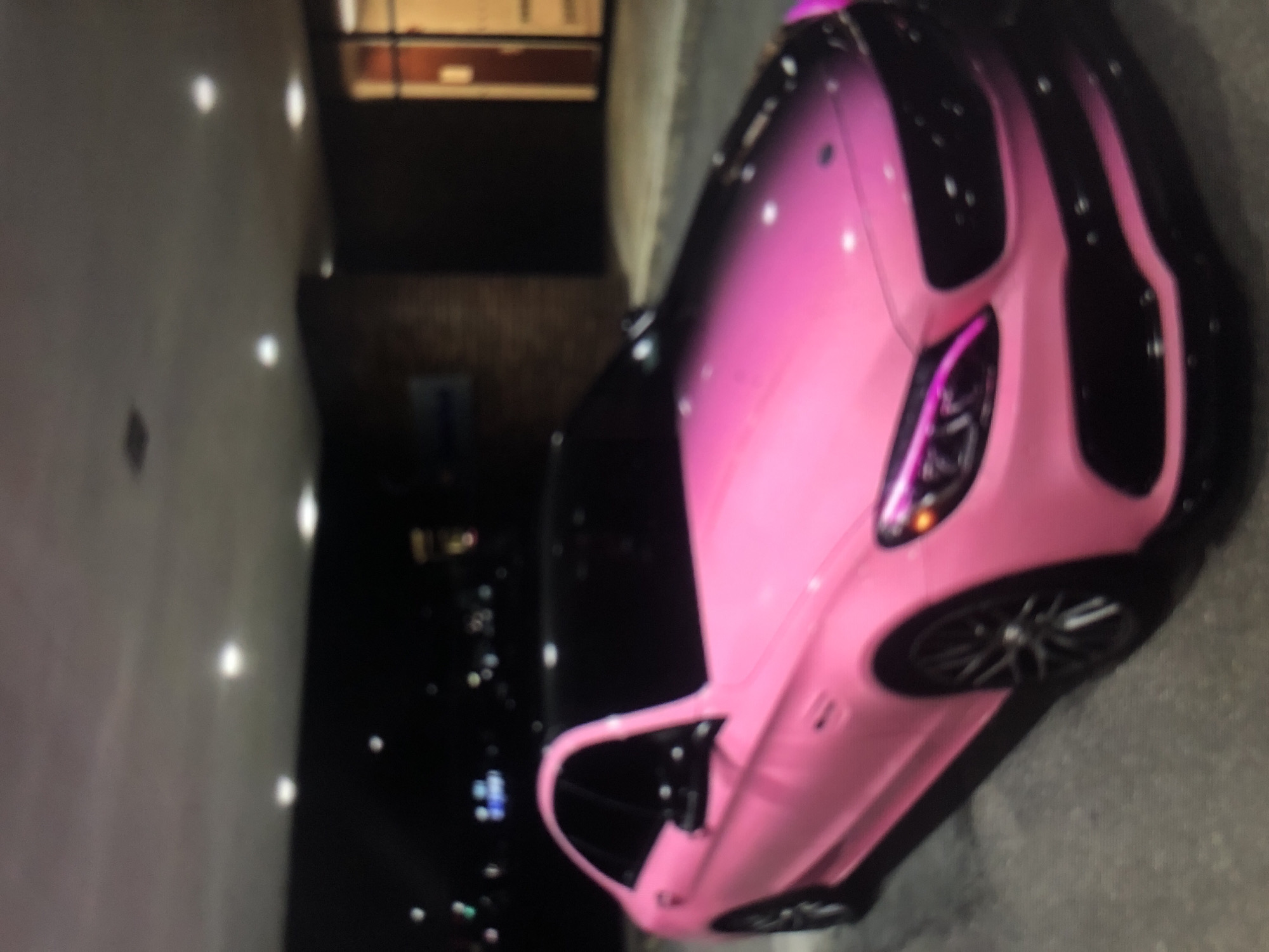 The image shows a pink car parked on a paved surface. A building is visible in the background. The location appears to be an urban area at night. The background features city lights.