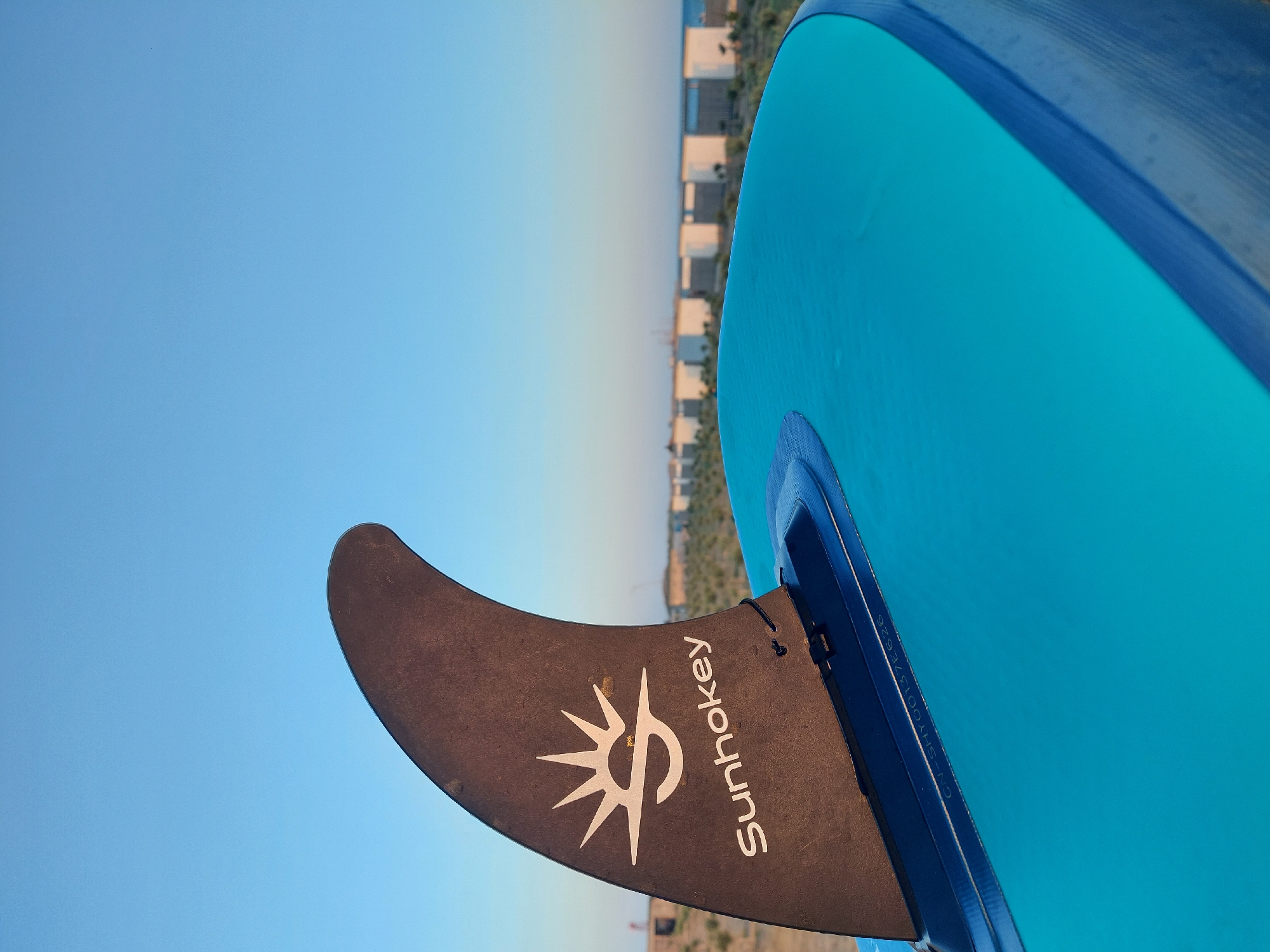 A close-up, eye-level shot focuses on the black fin of a blue stand-up paddleboard. The fin is angled upwards and to the left, with a white logo and the text "Sunhokey" printed on it. The fin is attached to the paddleboard with a blue mounting system. The background is a blur of pale blue sky, with a line of white buildings and green vegetation in the distance, hinting at a coastal location. The image appears to be taken outdoors in daylight.