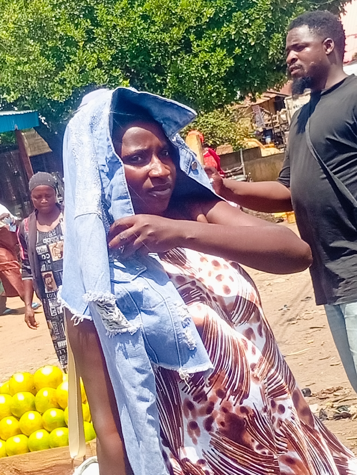 A medium shot captures a woman in Bukuru, Nigeria, partially obscuring her face with a light blue denim jacket draped over her head. She wears a patterned dress with brown and white abstract designs. To her right, a man in a black t-shirt and light blue jeans stands looking to his right, with his right arm extended towards the woman's head, appearing to adjust or hold the jacket. In the background, a stall with a large pile of bright yellow fruits, likely oranges, is visible to the left, with another woman in a patterned headscarf and dress standing behind it. Further back, market stalls and greenery are present. The ground is dusty.