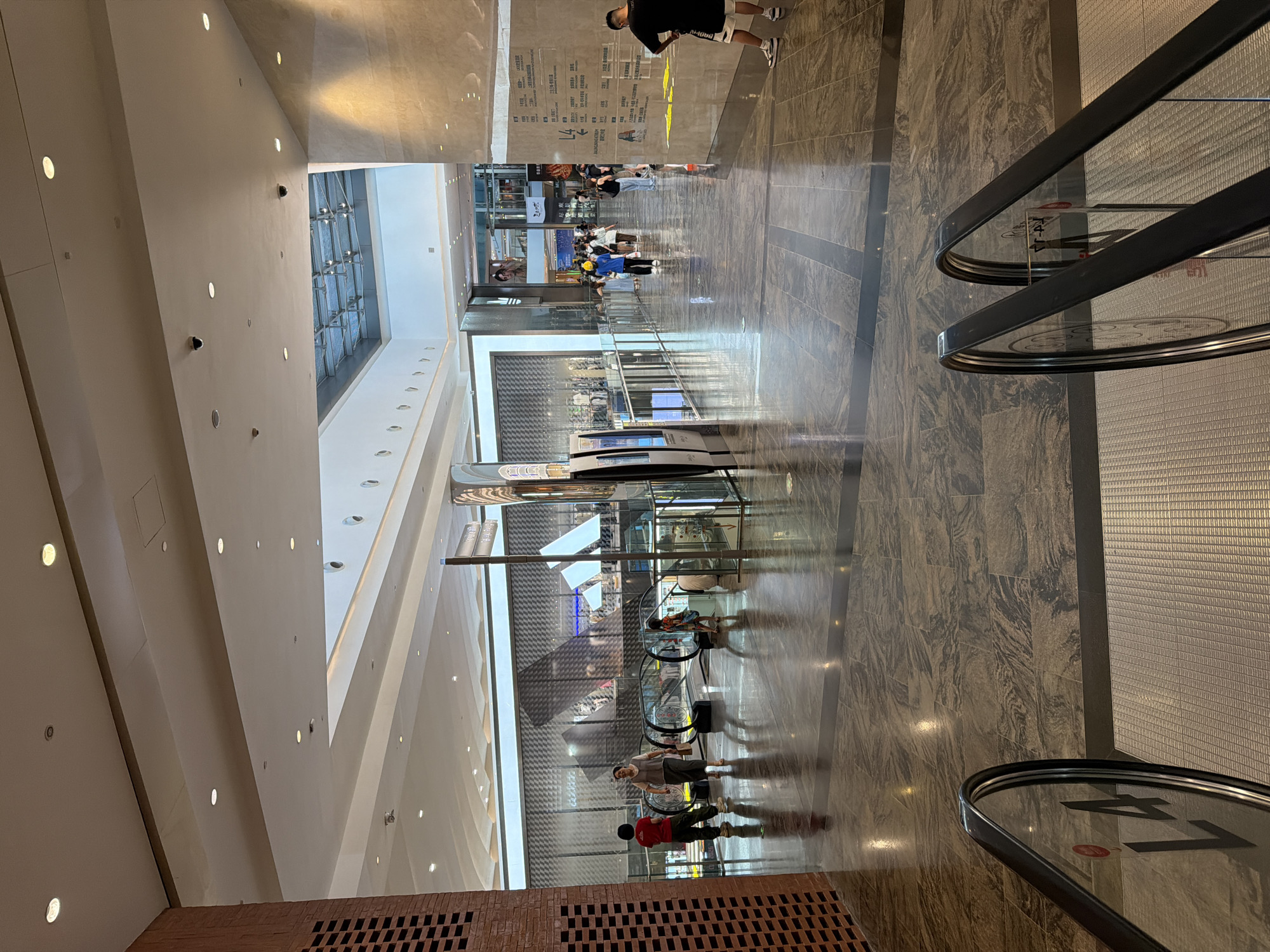The image shows a wide, marble-floored interior of a modern shopping mall in Yuzhong District, China. In the foreground, two escalators descend, with signage visible on the glass railings. A brick column stands on the left. In the middle ground, several people are walking, with two escalators going up behind them. A large, textured wall is visible on the right, adorned with directional signage. In the background, shops and displays are visible, and a glass-paneled ceiling allows natural light to enter the space. The overall atmosphere is bright and contemporary.