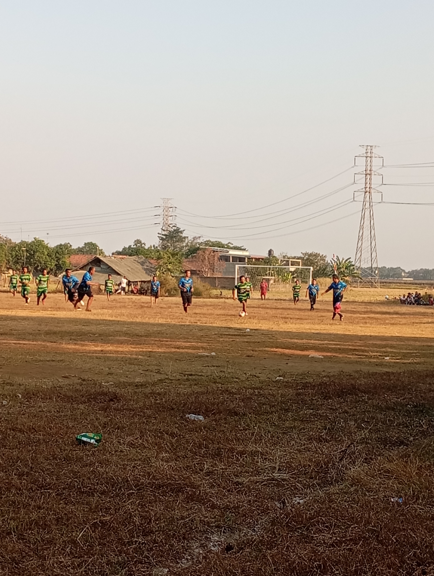 In Kecamatan Haurgeulis, Indonesia, a soccer game is in progress on a dry, grassy field under a pale sky. Several young males, wearing green and white striped jerseys or blue jerseys with black shorts, are actively playing. One player in a blue jersey is in mid-stride, appearing to kick a ball that is not visible in the frame. Another player in a similar blue jersey is running towards the left side of the frame, facing away from the viewer. In the background, a soccer goal with a white net is set up, and several spectators are seated on the ground to the right of the goal. Beyond the field, there are low-lying buildings with corrugated metal roofs and scattered trees. Two tall electrical towers with power lines stretching across the sky are prominent features of the landscape. The ground is covered in dry grass and some scattered litter, including a green and white chip bag and a plastic bottle.