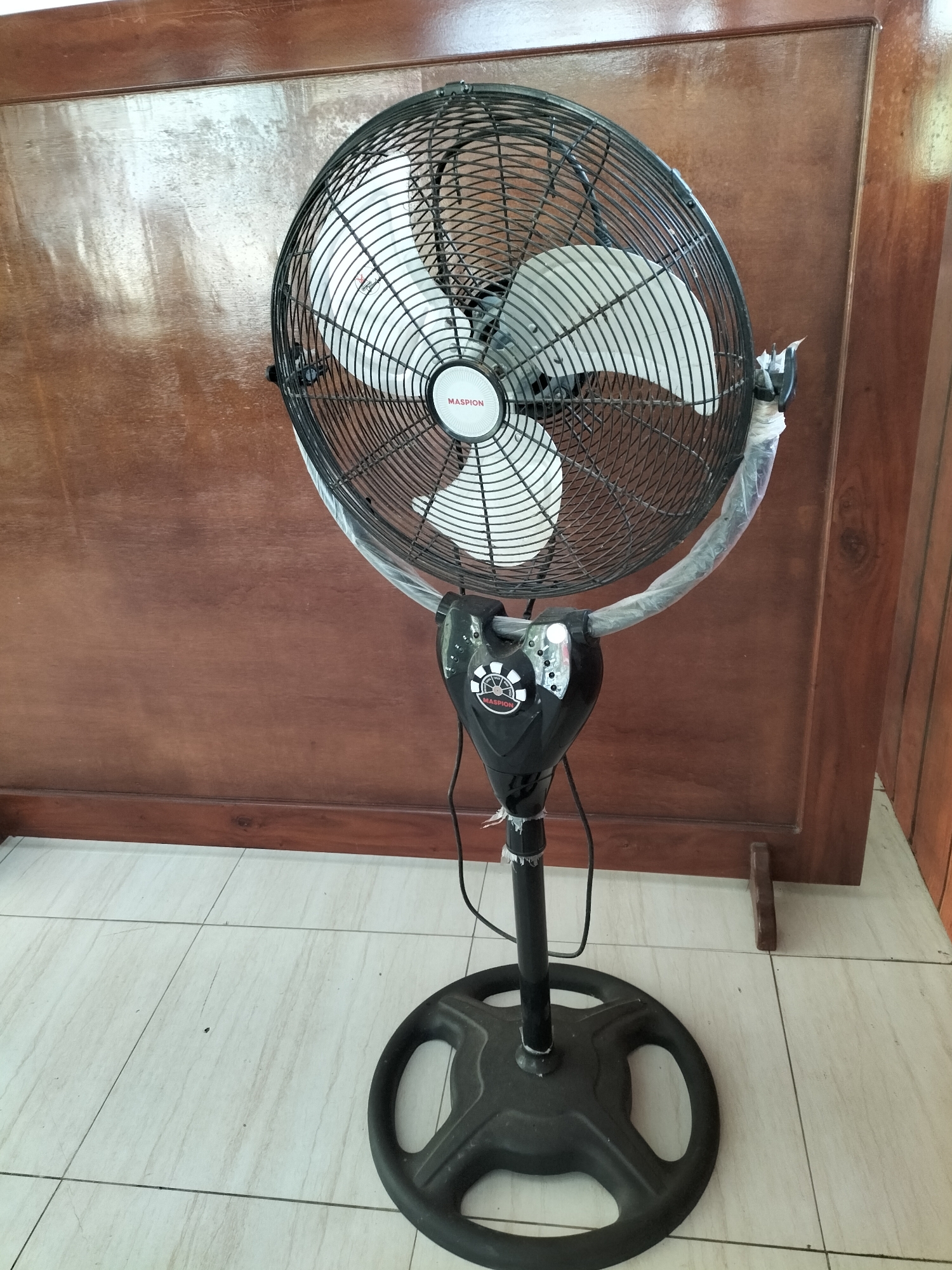 A black, oscillating pedestal fan with a white motor housing and clear plastic wrap around its neck stands on a white tiled floor in front of a dark wooden door. The fan's blades are white. The fan has a circular base and a black pole. The wooden door is smooth and polished, with visible grain. The scene is indoors, likely in a residential or commercial setting.