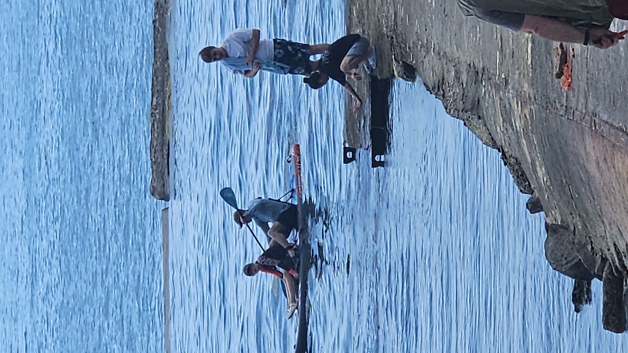 In a body of water that appears to be an ocean or large lake, two individuals are on a paddleboard. One person is seated and paddling, while the other sits in front. Adjacent to the water is a concrete structure, possibly a pier or seawall. On this structure, a person is crouched, reaching towards the water. Further along the structure, another person stands, holding a towel. The water is a deep blue, with visible ripples indicating movement. The concrete structure shows signs of wear and tear, with some debris present. The sky is not visible in the frame. The overall scene depicts a leisurely day by the water.