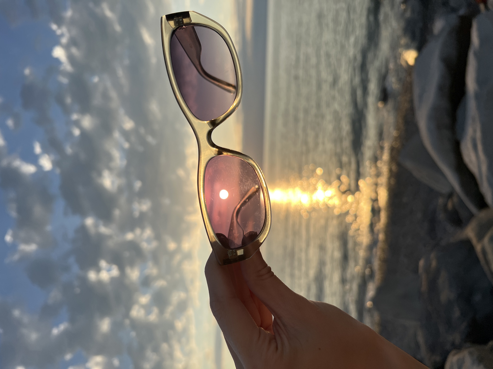 A person's hand holds a pair of sunglasses up to the sunset over the Black Sea. The sunglasses' lenses reflect the sun, creating a bright, circular glare. The sky is a mix of blue and white clouds, and the water is a deep blue with a shimmering reflection of the sun. The foreground is composed of large, grey rocks. The scene appears to be on a beach in Batumi, Georgia.