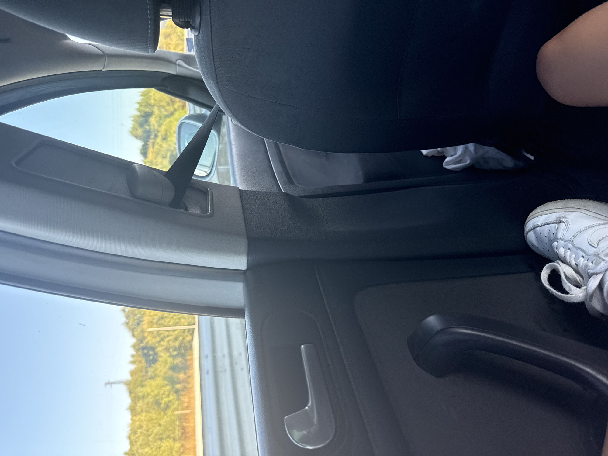 The image depicts the interior of a moving vehicle, looking out the right side window towards a sunlit landscape. The view from the window shows a blue sky with a few faint clouds, a power line tower on a distant, tree-covered hill, and a section of what appears to be a highway with a metal barrier. The interior of the car includes a grey seatbelt, a black car door with a silver handle, and the dark fabric of a car seat in the foreground. A white sneaker is visible on the floor of the car. The scene is consistent with travel on a road near Rudartsi, Bulgaria, given the environmental context.