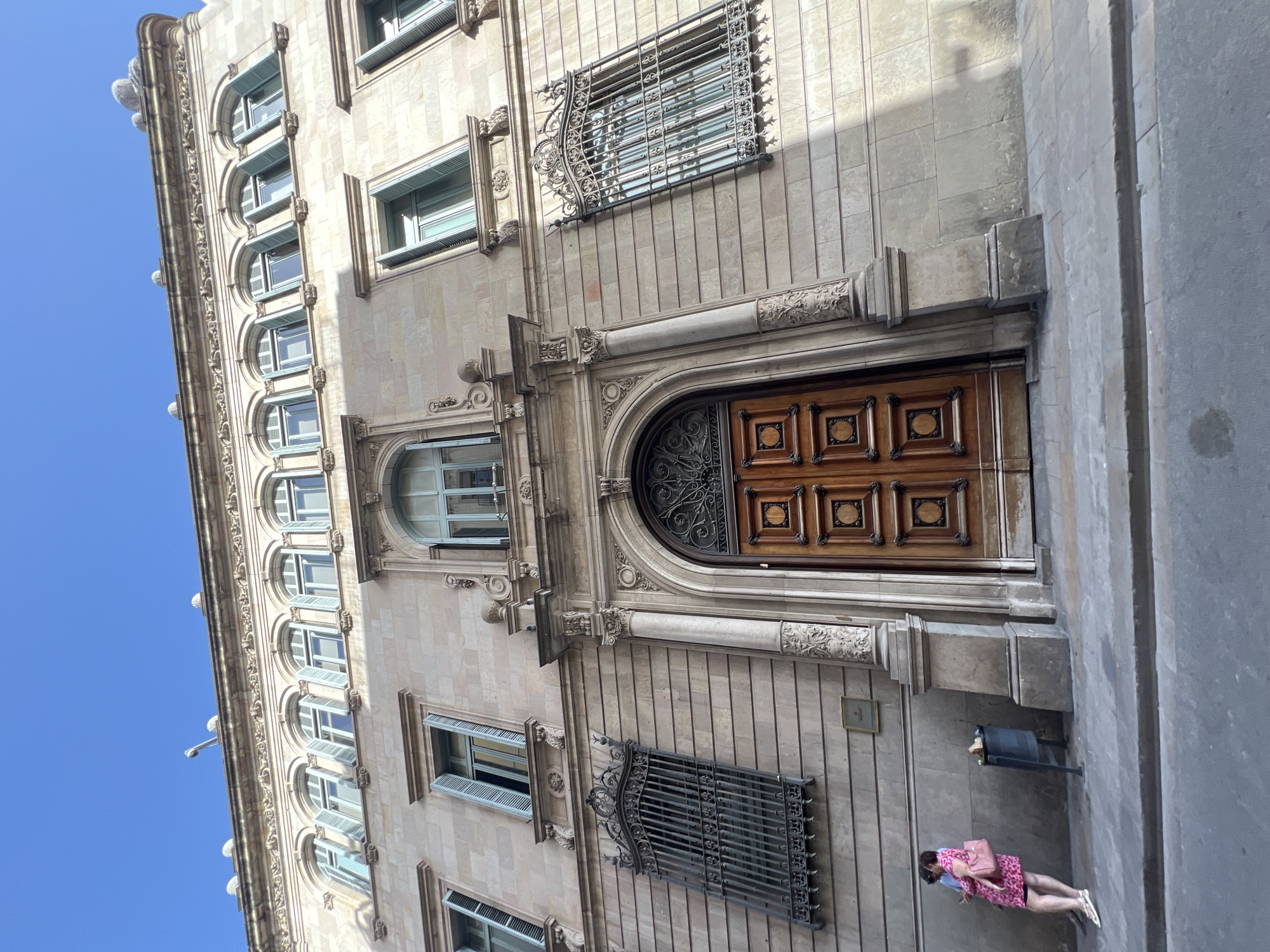 The image shows the facade of a building in Barcelona, Spain, with a wide wooden door and several windows. The building is made of stone and has ornate architectural details. A woman in a pink dress is standing on the sidewalk, looking at her phone. A trash can is next to her. The sky is clear and blue.