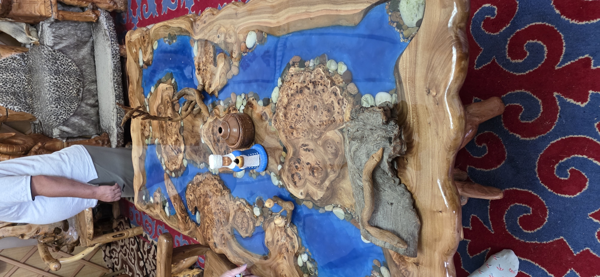 The image displays a unique, custom-made coffee table with a live-edge wooden top embedded with blue epoxy resin, resembling a river. The table is adorned with various natural elements such as stones, driftwood, and small decorative figurines. In the center of the table, a miniature wooden pot and a figurine of a person in traditional attire are placed. To the left of the table, a person wearing a white polo shirt and grey pants is partially visible, and to the right, a decorative wooden chair with animal print upholstery is present. The setting appears to be indoors, possibly in a residential space, as indicated by the patterned rug on the floor. The image does not provide any specific details about the location in Bulan-Segettu, Kyrgyzstan.