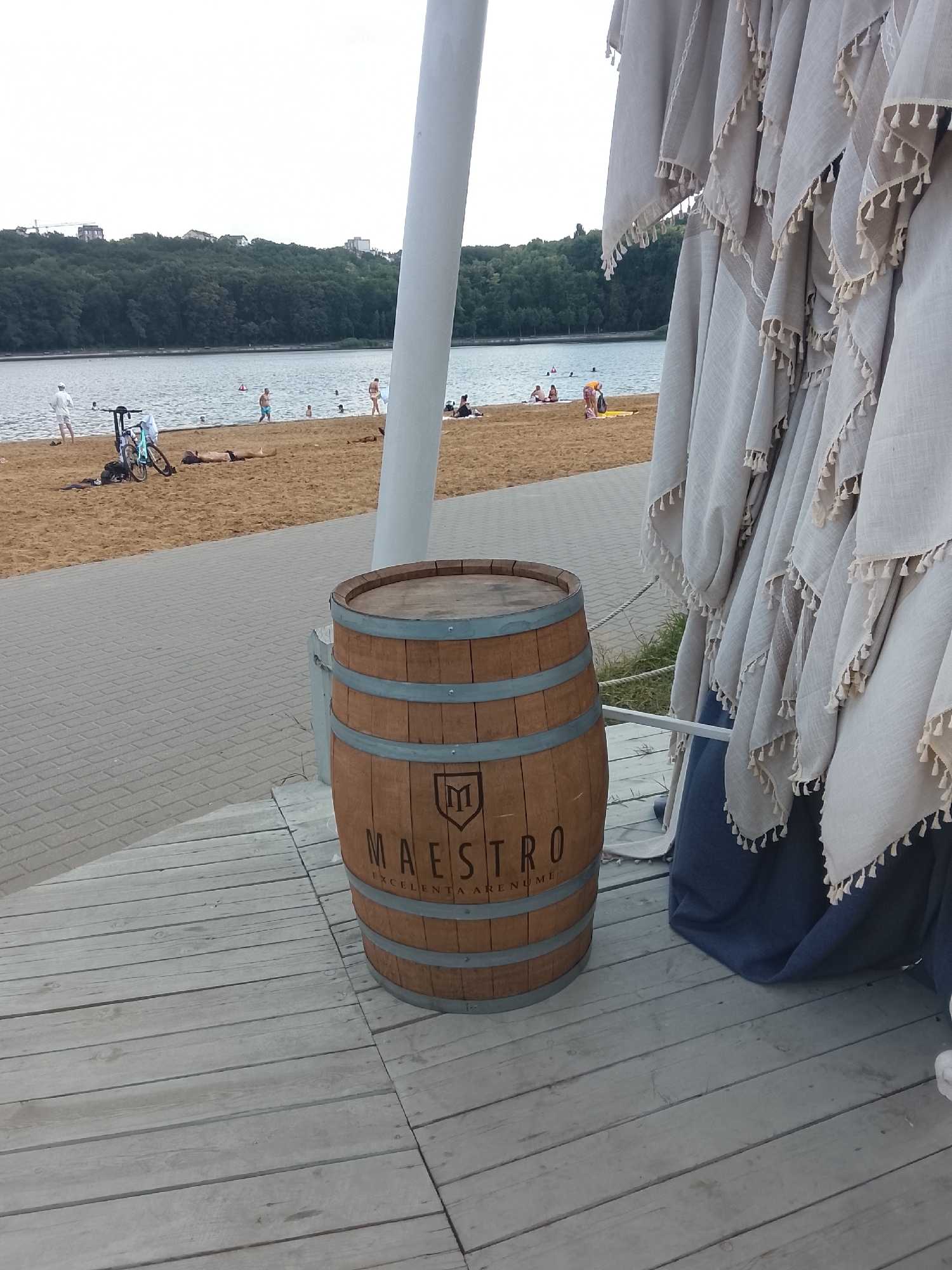 A wooden barrel with the logo "MAESTRO EXCELENTA ARENUME" is placed on a wooden deck next to a paved walkway. In the background, a sandy beach borders a body of water. Several people are visible on the beach, some swimming or sunbathing. A bicycle is parked on the sand near the walkway. On the right side of the image, several draped light-colored umbrellas with tassels are arranged. The sky is overcast. The scene appears to be a recreational area, possibly a beach or waterfront park.