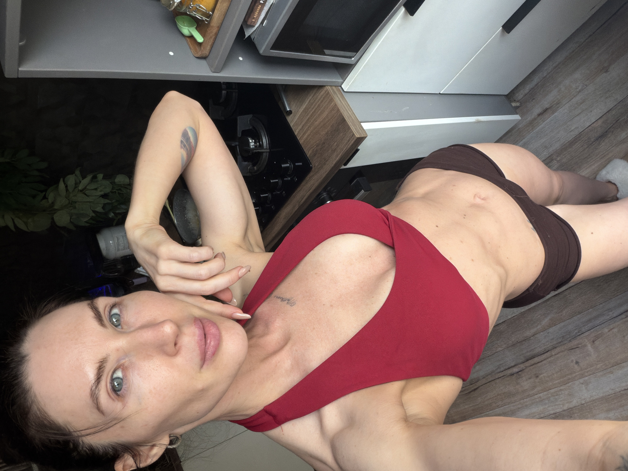 A woman is depicted taking a selfie while lying down, with the image rotated 90 degrees clockwise. She is positioned on a light wood-planked floor in what appears to be a kitchen.
The woman is wearing a red sports bra or crop top and dark brown shorts or underwear, showcasing a lean physique. She has light-colored eyes, dark hair, and light skin, with a neutral expression as she looks directly at the camera. Visible tattoos include a multi-colored, feather-like design on her left bicep and script text on her upper left chest, which appears to read "Mother." One foot, clad in a white fuzzy slipper, is visible at the bottom right of the frame.
The background reveals typical kitchen elements. A dark-colored gas stovetop with black control knobs is seen under light grey and white kitchen cabinets. A microwave oven is built into the cabinetry, displaying a label with partial text "БЕДНОВА Р." Above the stovetop, a wooden cutting board holds a clear jar and a green measuring spoon. Dark foliage or plants are visible in the lower left background. The overall scene suggests an indoor setting in Perm, Russia.