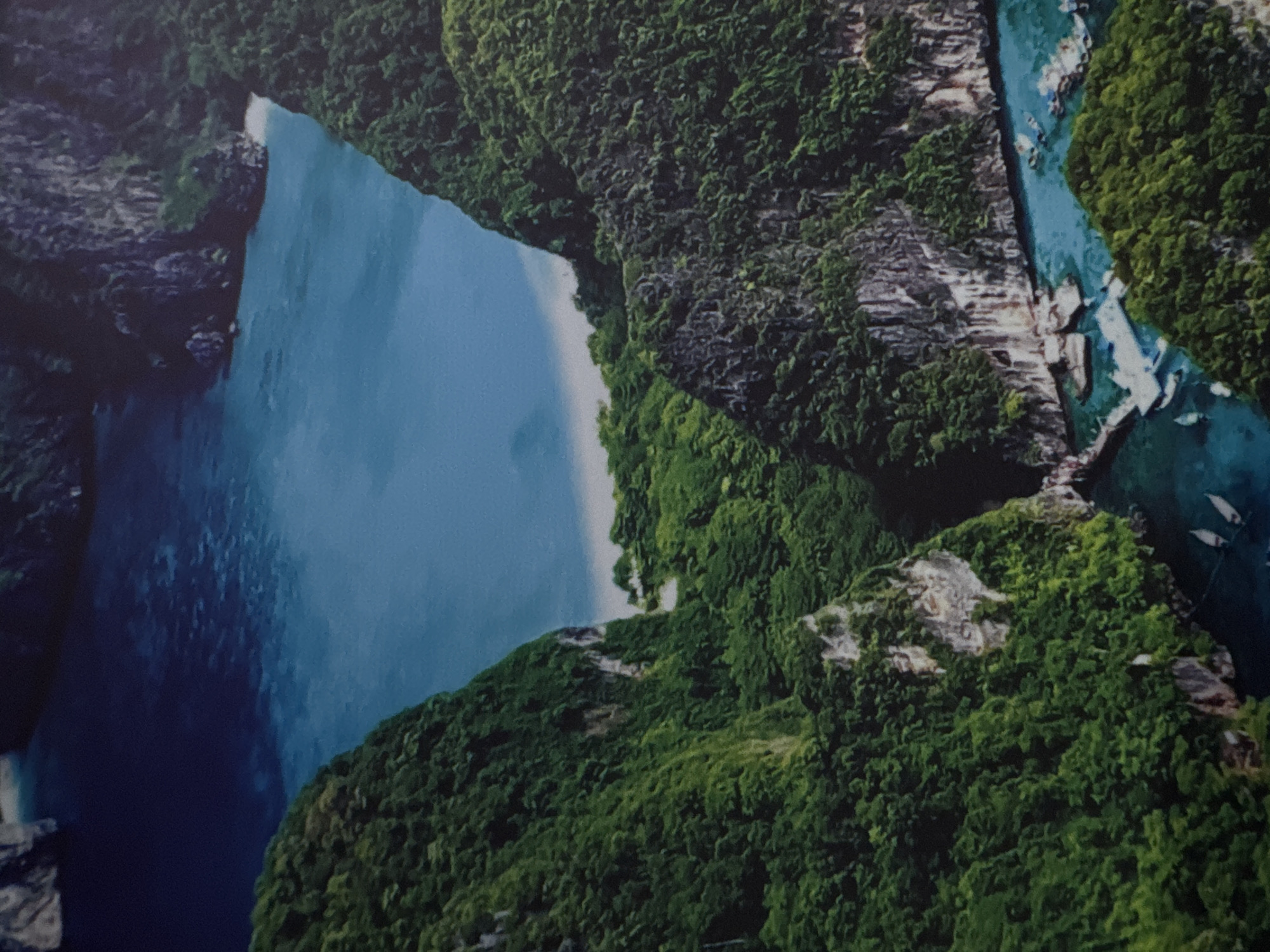 This is an aerial, sideways photograph of a tropical island with lush green foliage covering steep, rocky cliffs. A calm, turquoise body of water surrounds the island, and a small beach is visible on one of the shores. Several white boats, likely traditional longtail boats, are moored in the water. The scene is bathed in natural light, suggesting it is daytime. The image does not contain any visible text. The overall impression is one of natural beauty and tranquility.