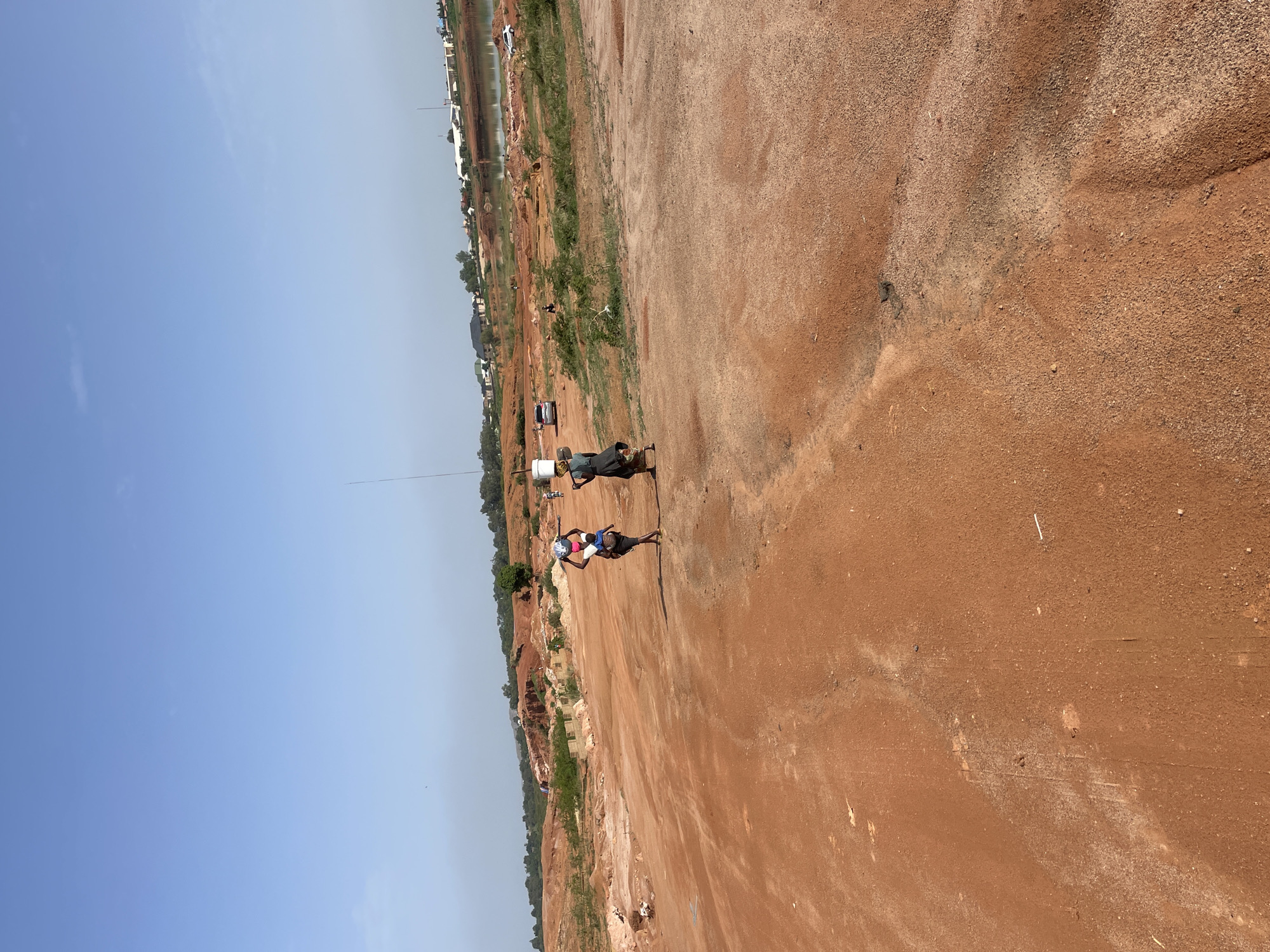 The image depicts an open, red-earth expanse in Jos, Nigeria. A few buildings are visible in the background, suggesting a peri-urban setting. In the foreground, two people are walking. One is wearing a blue shirt and carrying something on their head, and the other is wearing dark clothes and carrying a container on their head. The ground is bare and reddish in color, transitioning to a mix of grass and scrub vegetation closer to the buildings. The sky is clear and blue. A cell tower is visible in the distance.