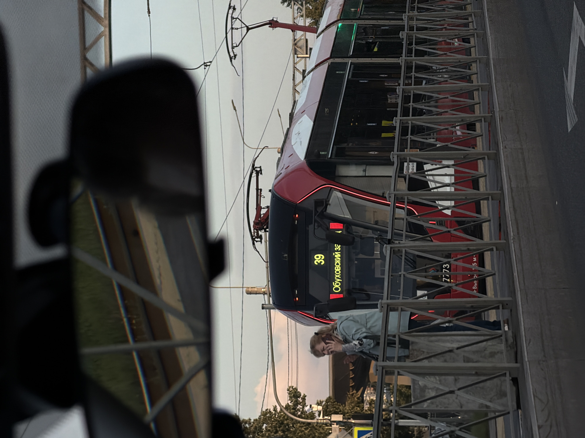 The image captures a scene in Saint Petersburg, Russia, likely from inside a vehicle, as evidenced by the reflection in a rearview mirror in the foreground. A modern, red and white tram, numbered 39, is stopped at a station. The tram displays "Обуховский за" on its digital sign, indicating its route. To the left of the tram, a woman with blonde hair is standing on the platform, holding a mobile phone to her ear and appearing to be engaged in a conversation. She is wearing a light blue jacket and dark pants. A metal railing separates the tram platform from the sidewalk. The environment suggests an urban setting with overhead power lines for the tram and some trees visible in the background. The sky is overcast with hints of pinkish clouds, indicating it might be dusk or dawn. The overall scene is a candid snapshot of daily urban life.