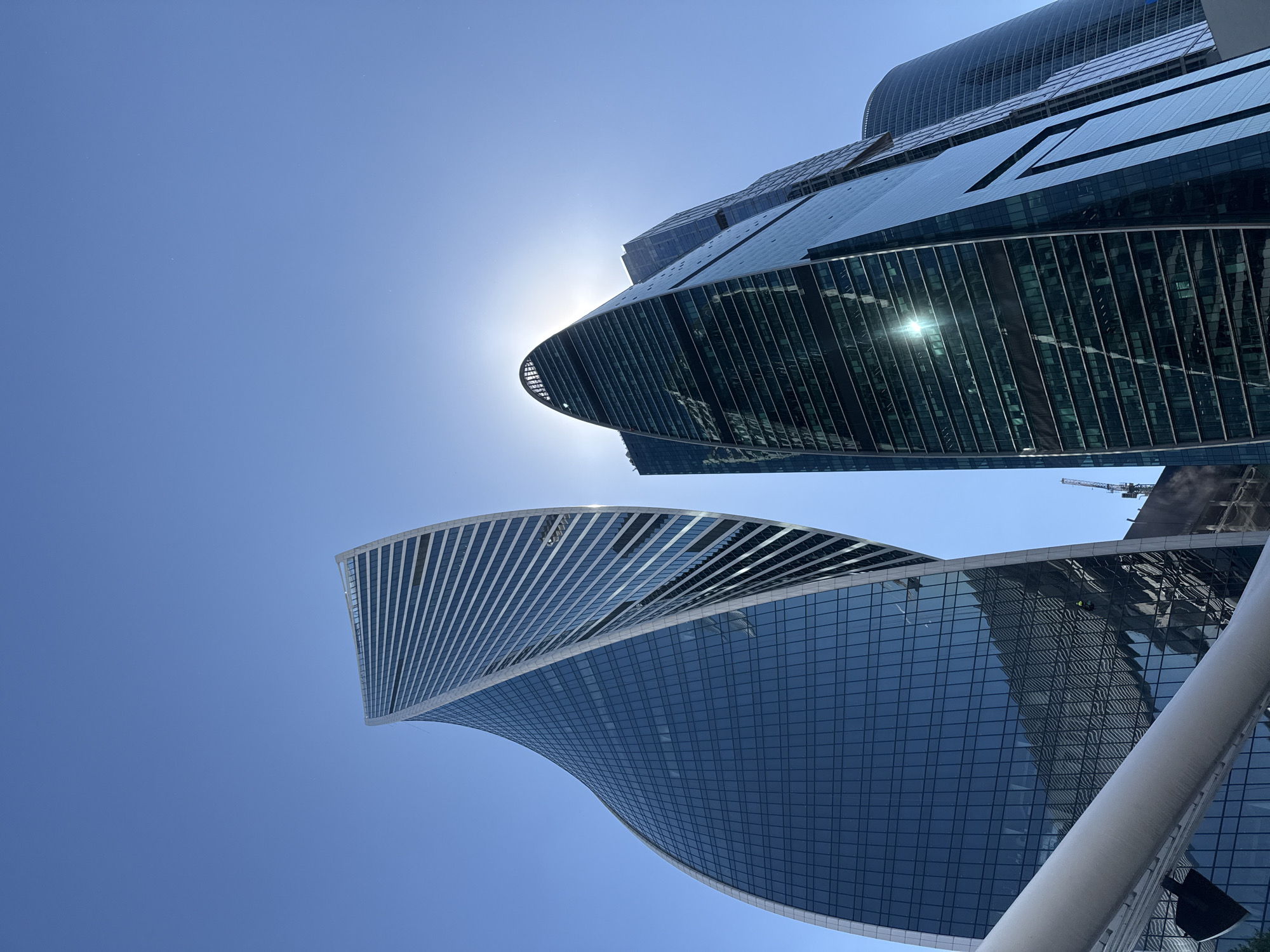 This image displays a low-angle perspective of modern skyscrapers in the Moscow International Business Center (Moscow-City), Moscow, Russia, under a clear blue sky. The prominent structure on the left is the Evolution Tower, characterized by its distinctive helical, twisting glass facade, featuring dark blue windows and white spiral framework. A window cleaner wearing a bright yellow/green safety vest is discernible rappelling down the Evolution Tower's facade in the lower-mid left section. To the right, a second skyscraper, identified as part of the IQ-quarter complex, exhibits a pointed, curved apex. The sun is positioned directly behind this building, generating a powerful silhouette, lens flare, and a bright reflection on its lower glass facade. A third, partially visible cylindrical skyscraper is present in the far right background. A substantial, light-colored, tubular architectural element diagonally crosses the lower foreground from left to right. Minor hints of construction activity, including a crane, are visible in the mid-ground between the structures. The sky is uniformly bright blue, indicating clear weather.