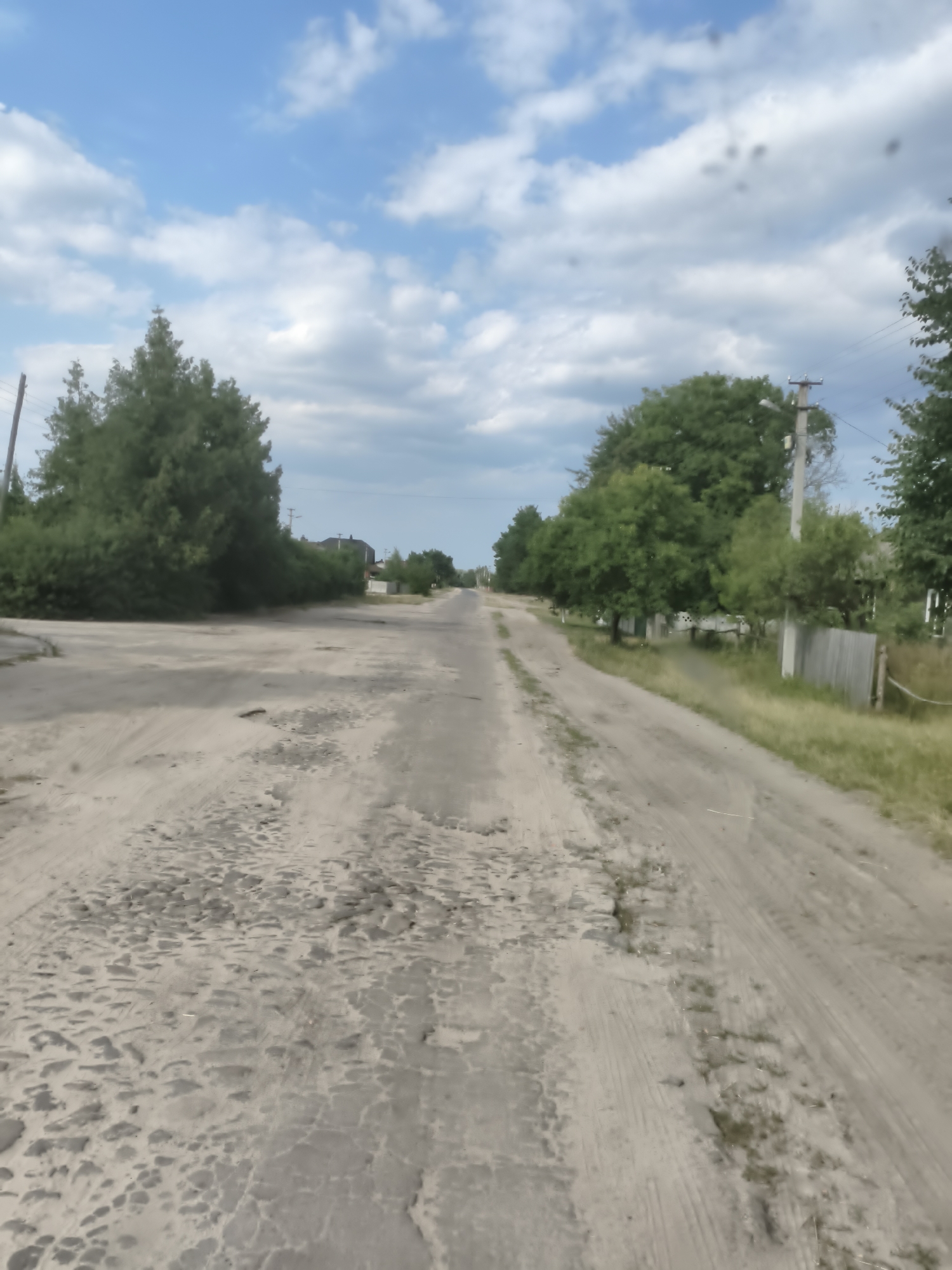 The image depicts a dirt road in the city of Bohushi, Ukraine, under a partly cloudy blue sky. The road is heavily potholed and appears to be in disrepair, with loose gravel and dirt. Trees line both sides of the road, with dense foliage on the left and a mix of trees and fences on the right. In the distance, houses and utility poles are visible, suggesting a rural or semi-rural residential area. The overall scene conveys a sense of an unpaved, weathered rural landscape.