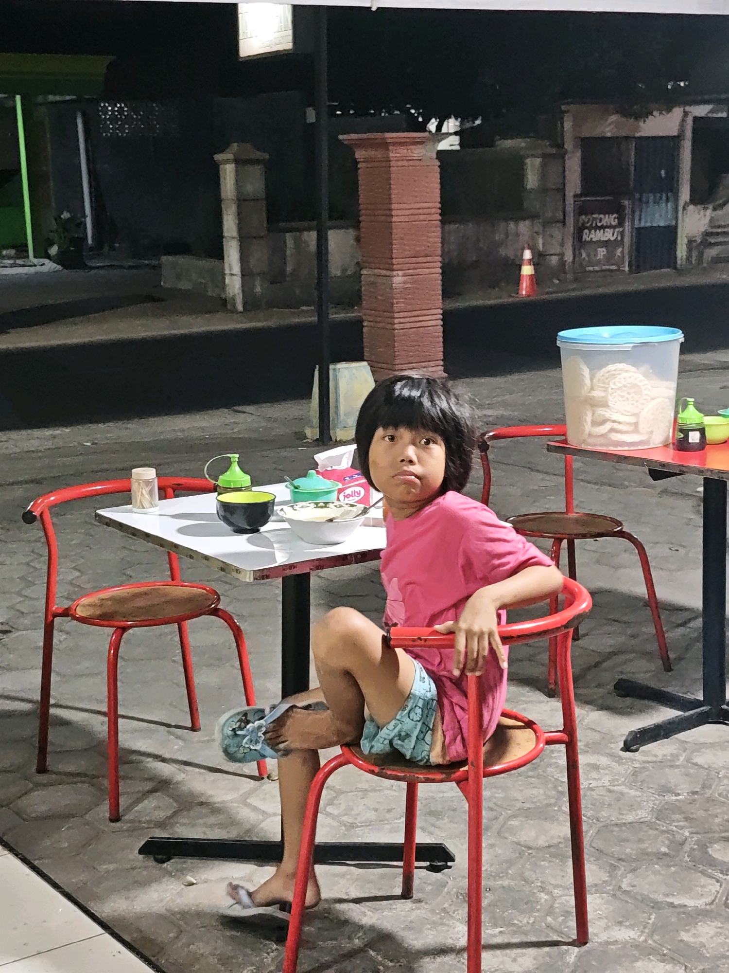 In Ngasem, Indonesia, a young child with dark hair and a serious expression sits at a white table in front of a red chair. The child wears a pink t-shirt and light blue shorts with a cartoon pattern. The child's legs are crossed, with one foot bare and the other wearing a light blue sandal. On the table are several items, including a black and green bowl, a small green bottle with a spout, a small container with toothpicks, and a box of tissues. Another red chair is visible to the left of the table, and a second table with a container of crackers is in the background. The scene appears to be outdoors, with a paved ground surface. In the background, a building with a sign that reads "POTONG RAMBUT JP" and a traffic cone are visible. The lighting suggests it is nighttime or late evening.